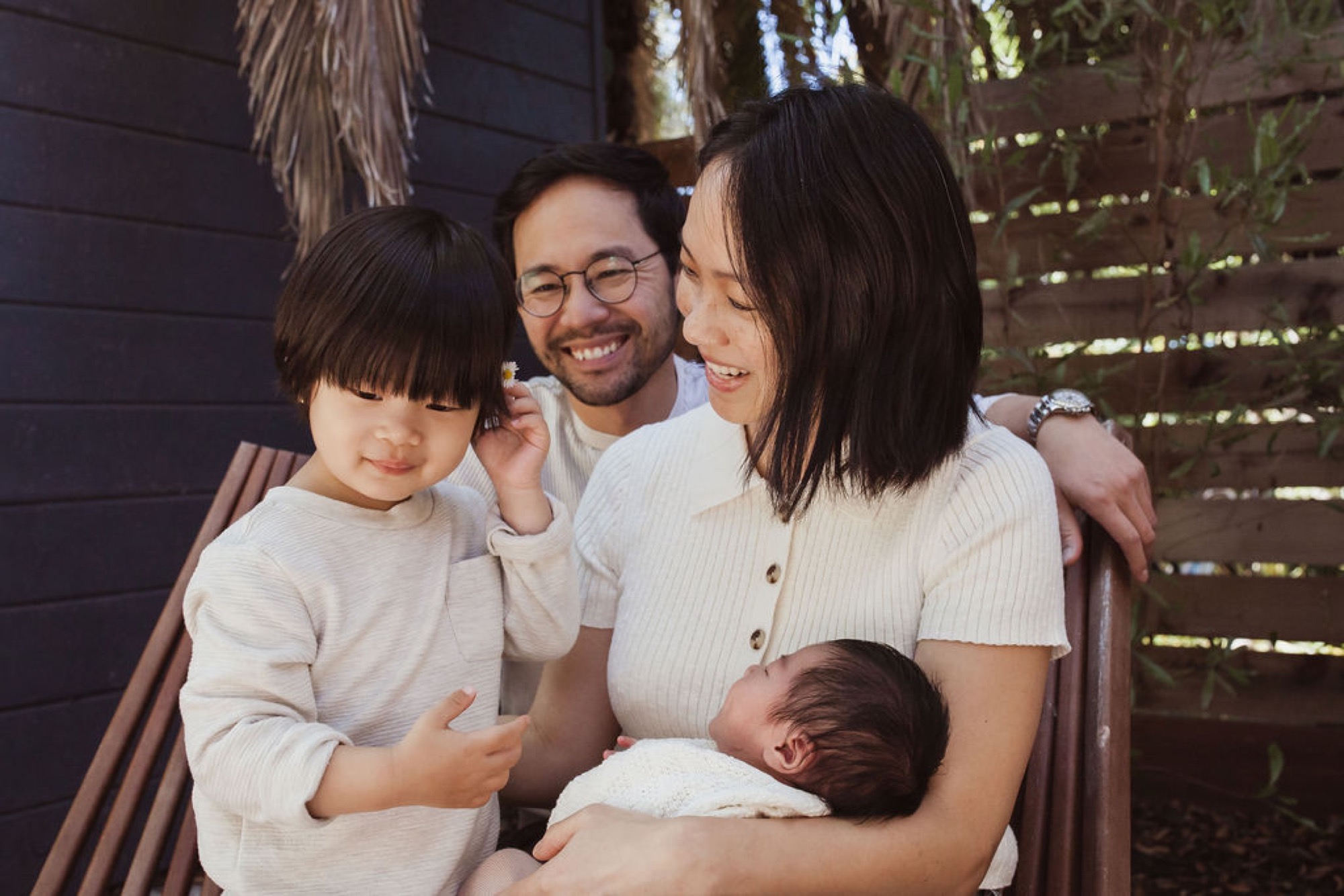Young family of four shares genuine laughter in dappled shade, with mother cradling newborn while toddler and father beam beside them
