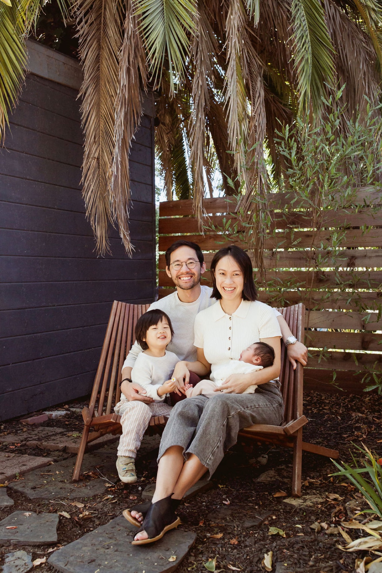 Family of four sits together on a wooden chair beneath hanging palm fronds, all beaming with genuine joy and warmth