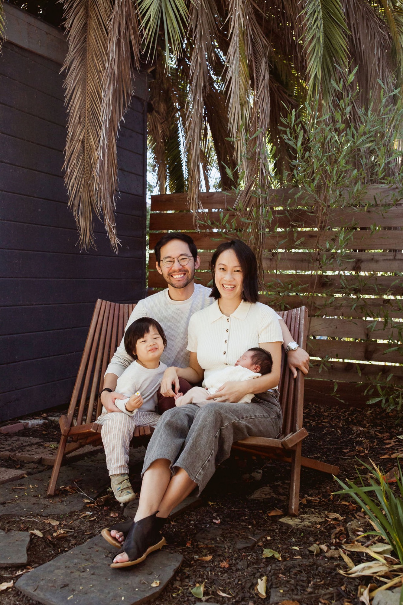 A family of four sits together on a wooden bench beneath hanging palm fronds, all smiling warmly at the camera in dappled afternoon light