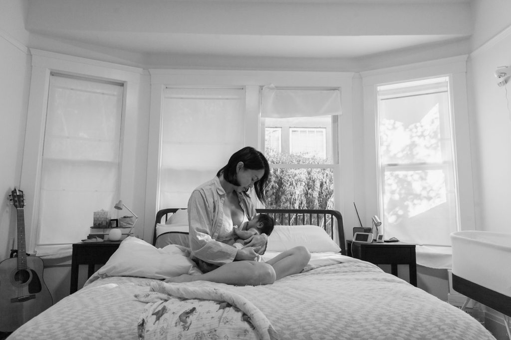 Mother gazes down at newborn cradled in her lap on a sunlit bed, soft natural light streaming through bay windows behind them