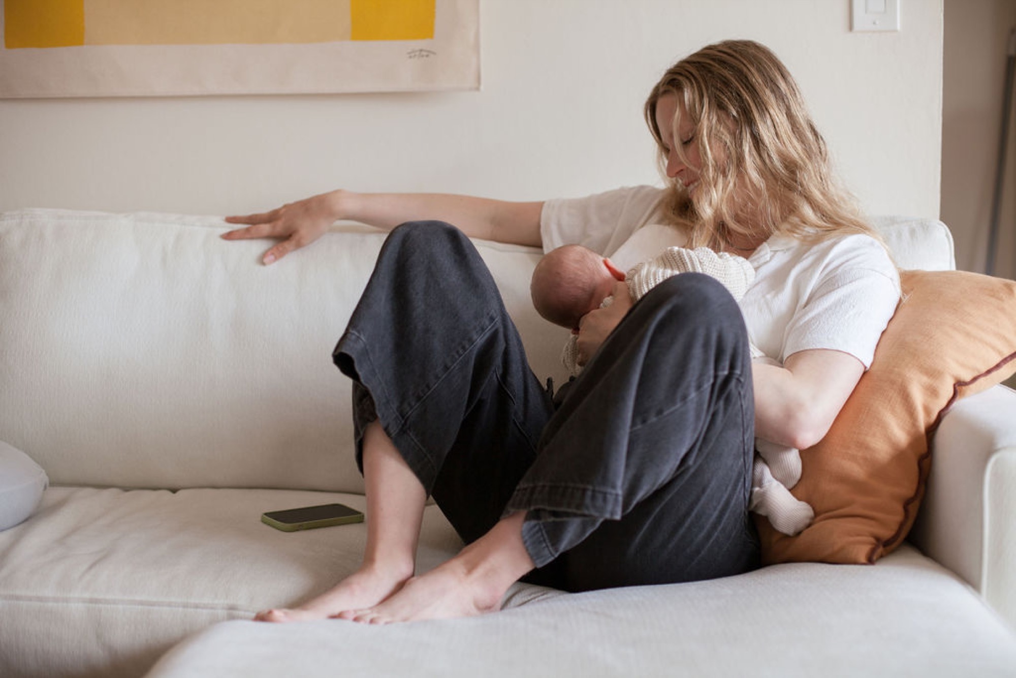 Mother relaxes on a cream sofa, cradling her newborn wrapped in white while gazing down with tender affection