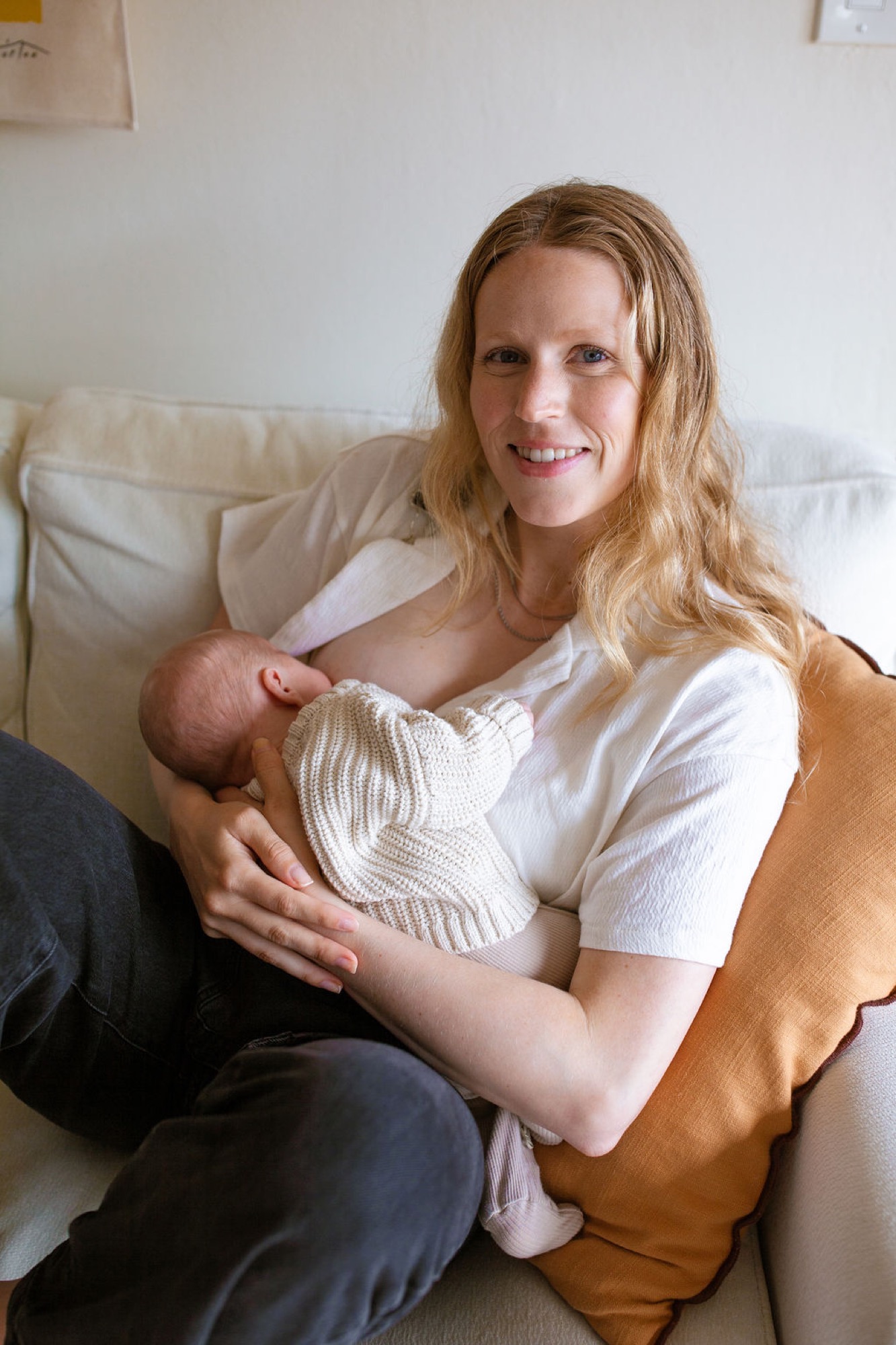 Mother in mustard cardigan smiles warmly while cradling newborn in cream wrap on neutral couch, soft natural light illuminating her gentle expression