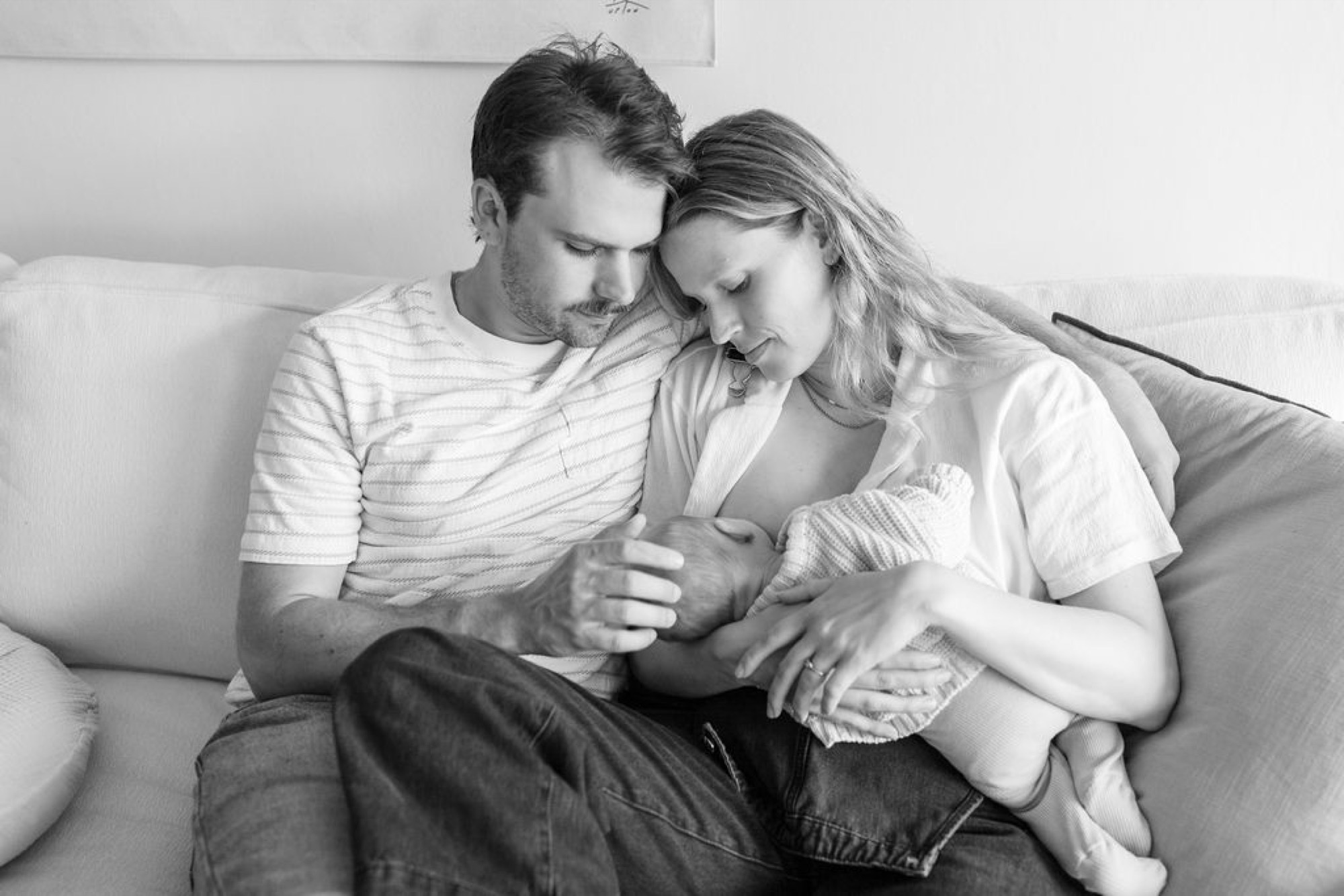 Parents gaze tenderly at their newborn on a bright couch, soft natural light illuminating their gentle expressions and intimate connection