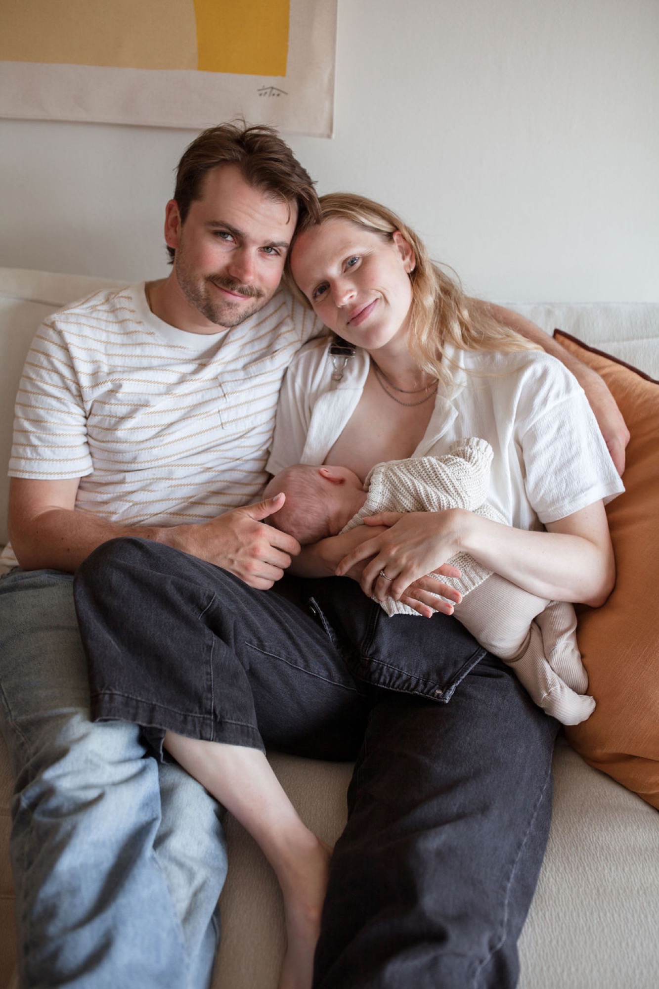 New parents sit closely on a neutral sofa, tenderly cradling their swaddled newborn between them with warm, genuine smiles