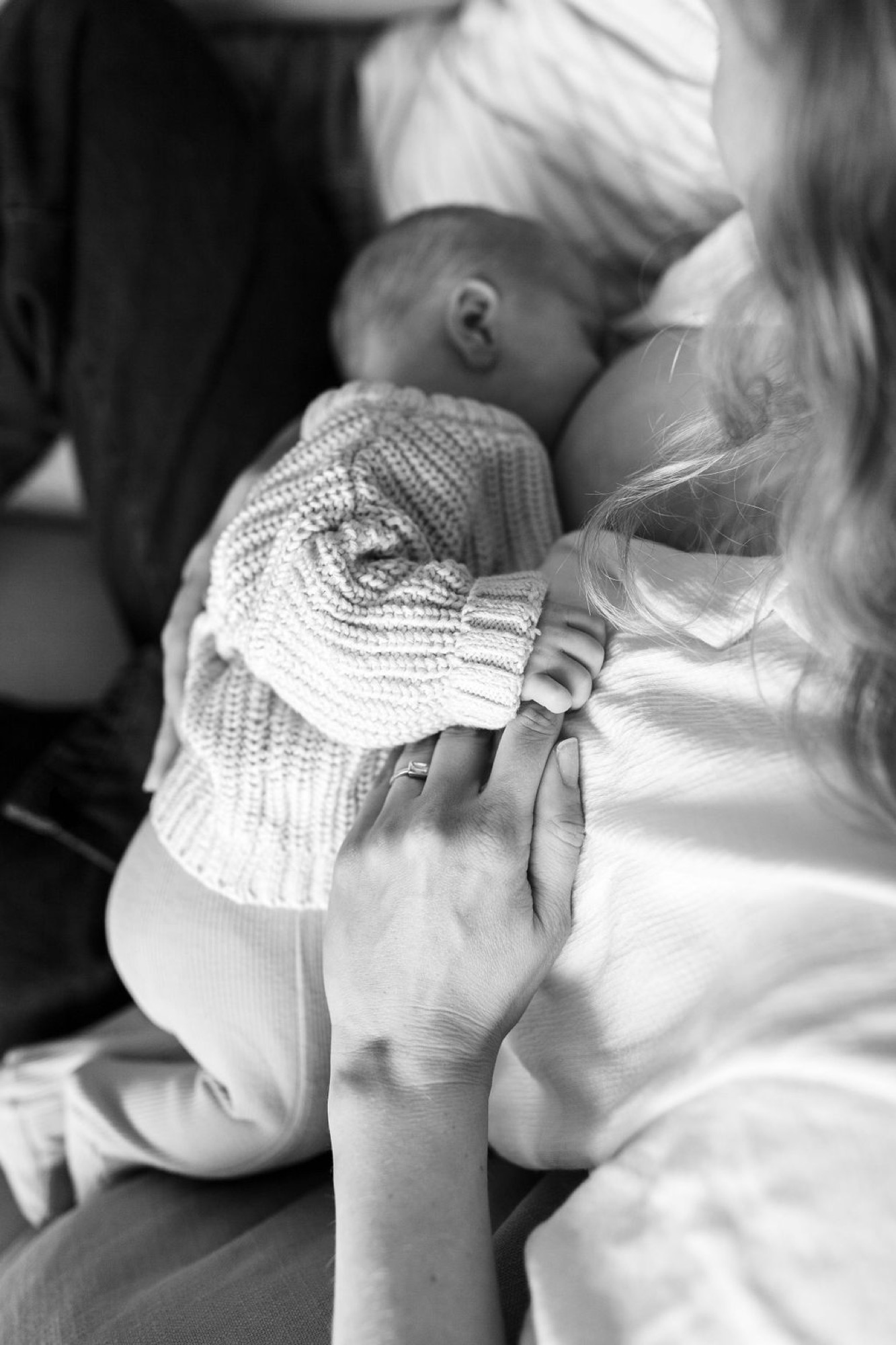 A newborn swaddled in a knit blanket nestles against their mother's chest, tiny hand grasping fabric while she gazes down with tender affection