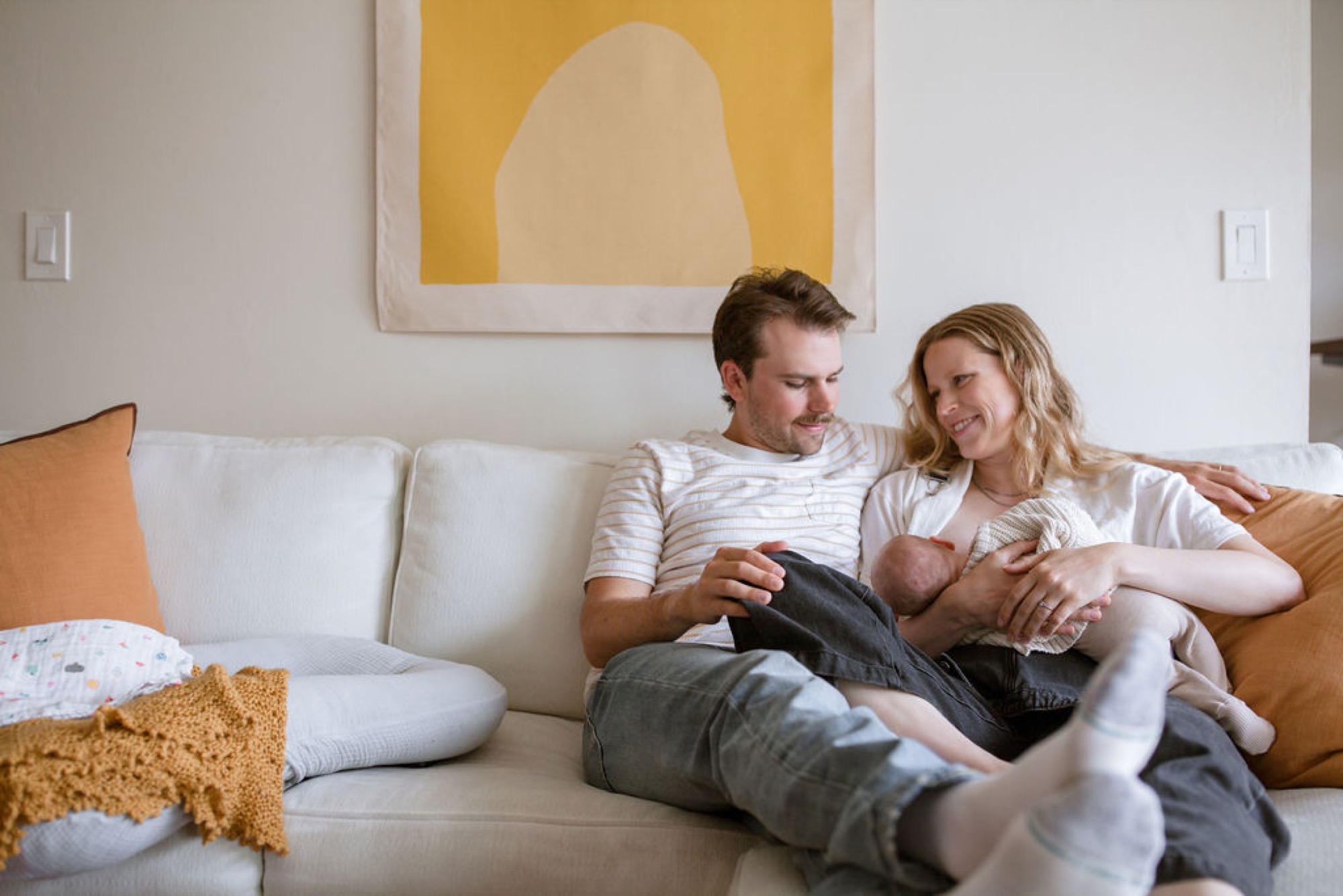 New parents sit together on a cream sofa, the father smiling down at his newborn while the mother gazes at her infant with tender affection