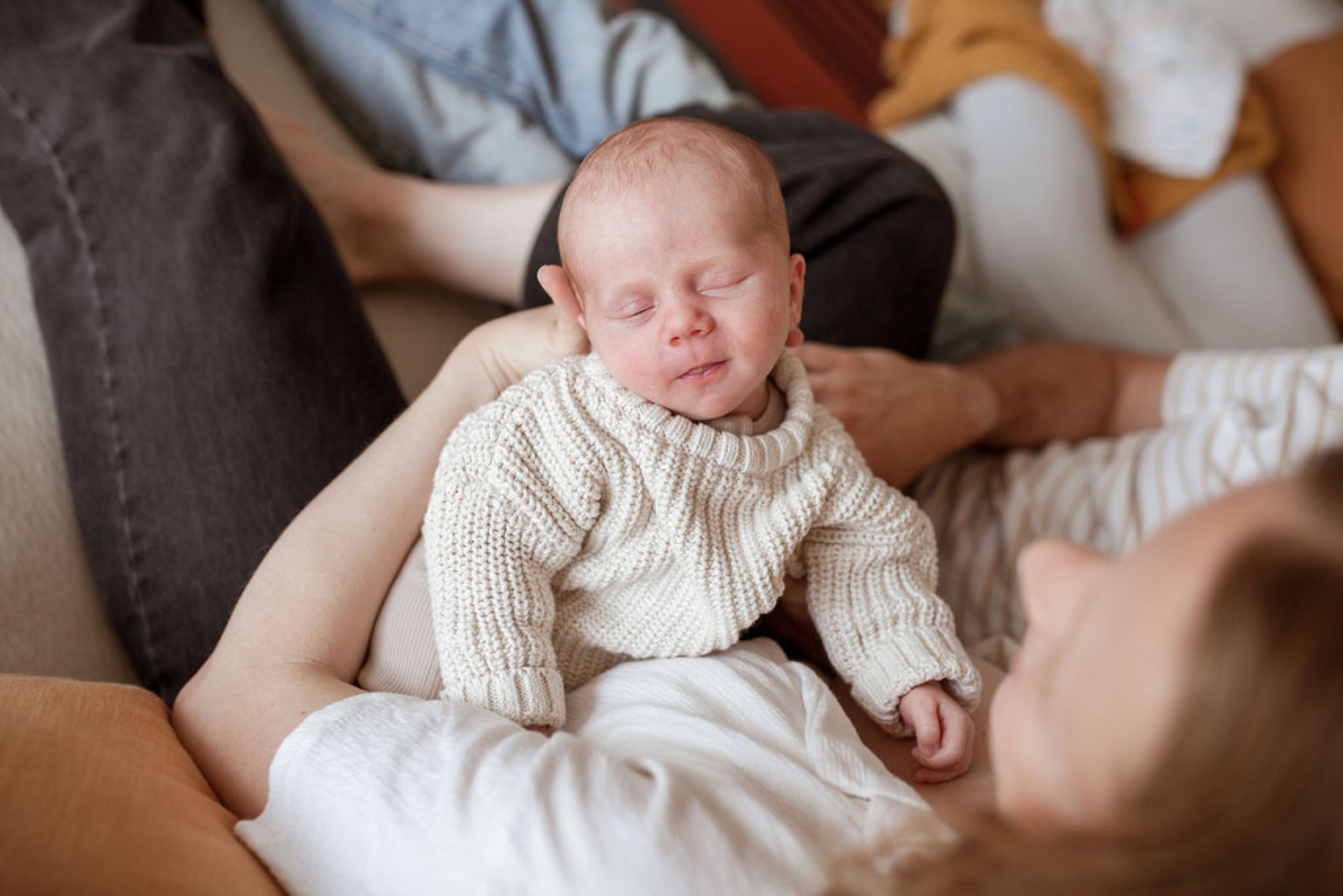 A peaceful newborn sleeps peacefully in a cream knit sweater, cradled by a parent's hands with family gathered closely around on a couch