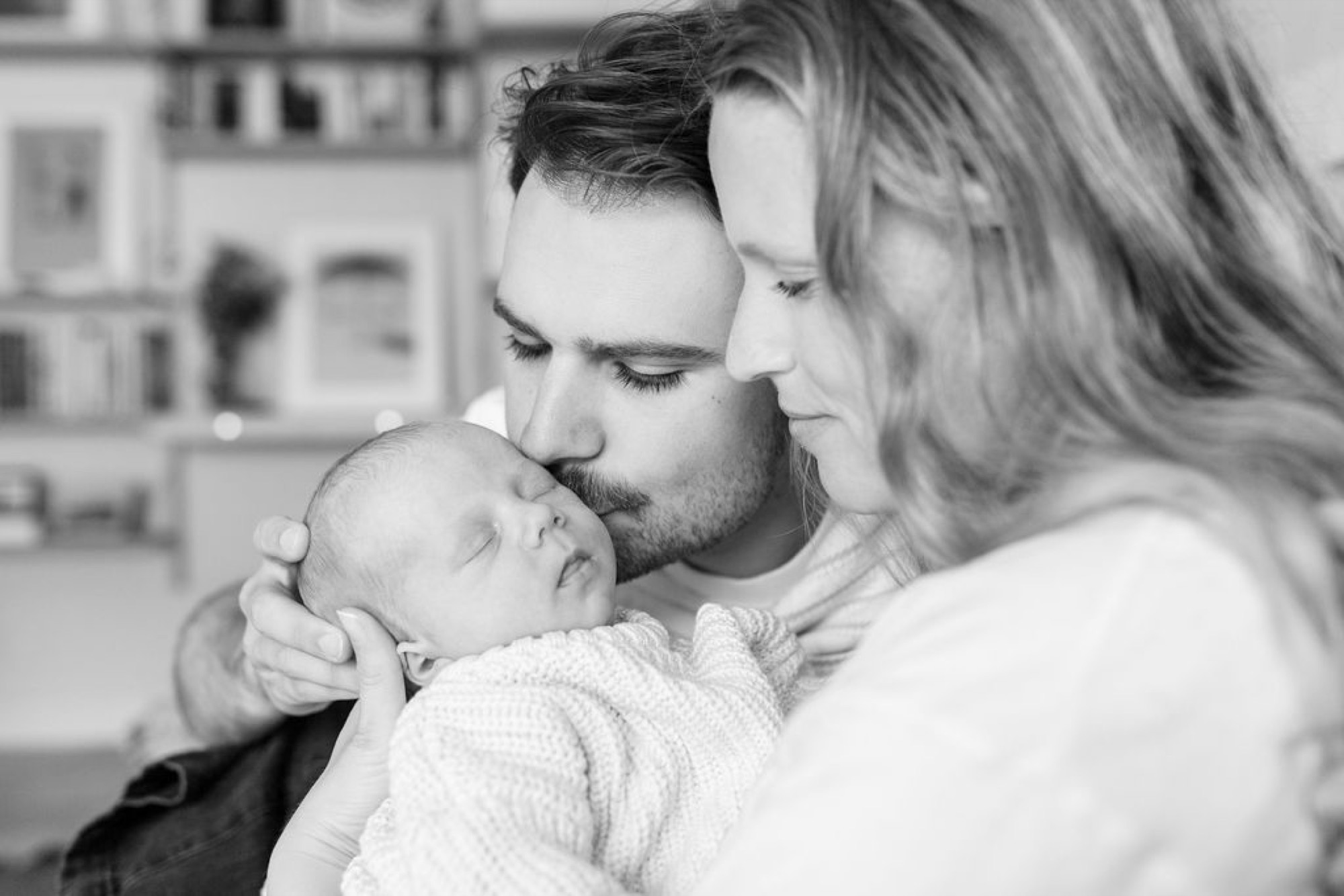 A bearded father tenderly kisses his newborn's forehead while the mother gazes down with soft affection in their bright, minimalist home
