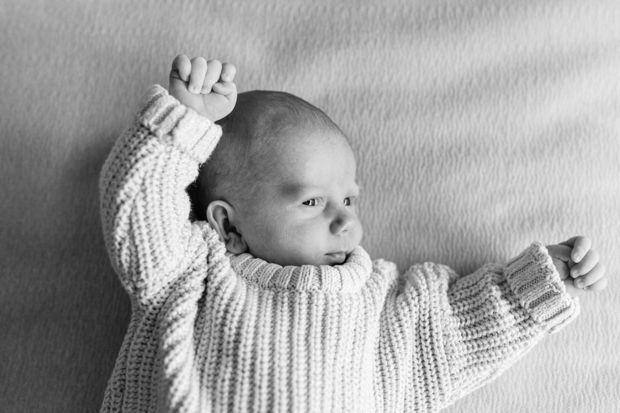 Newborn in cream cable-knit sweater stretches both arms overhead against textured white wall, head turned in profile with alert expression