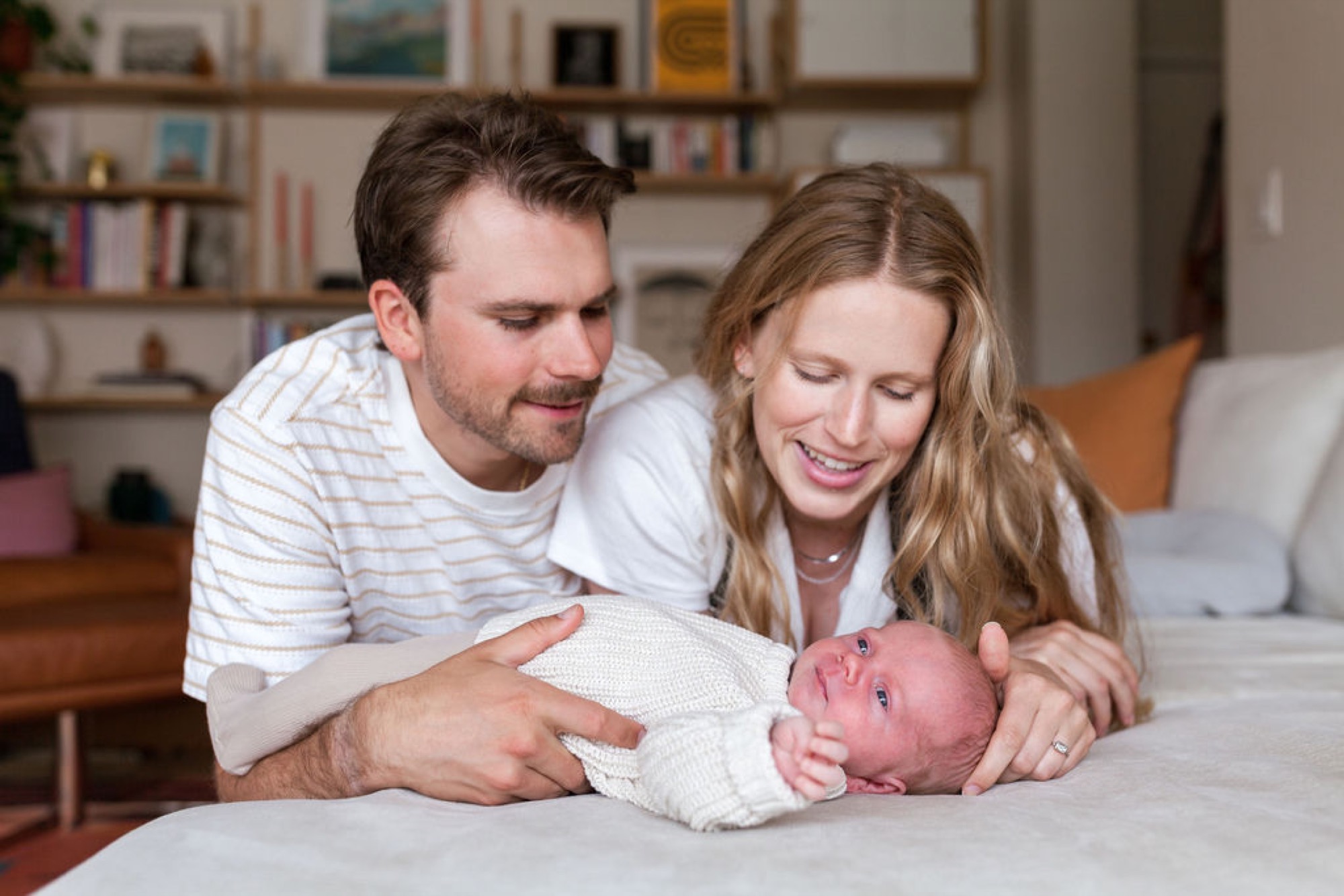 Parents beam down at their newborn wrapped in cream knit on a bed, soft natural light illuminating their joyful expressions and tender connection