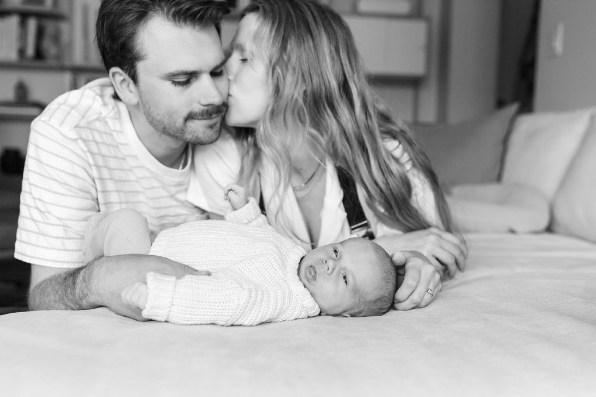 Parents gaze tenderly at their newborn swaddled in white on a soft bed, captured in intimate black and white portraiture