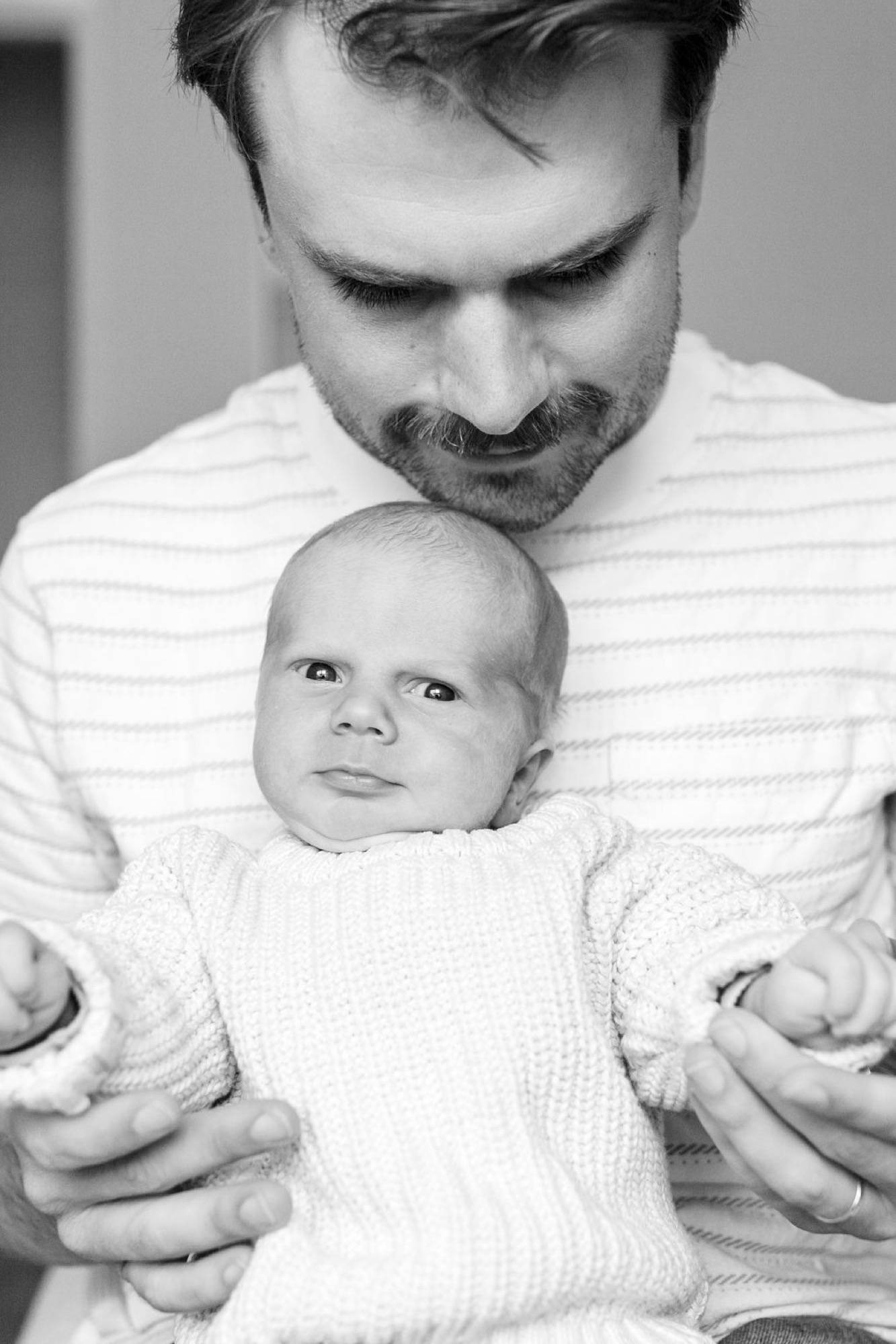 A bearded father gazes tenderly downward at his swaddled newborn, who stares directly at the camera with alert, wide eyes