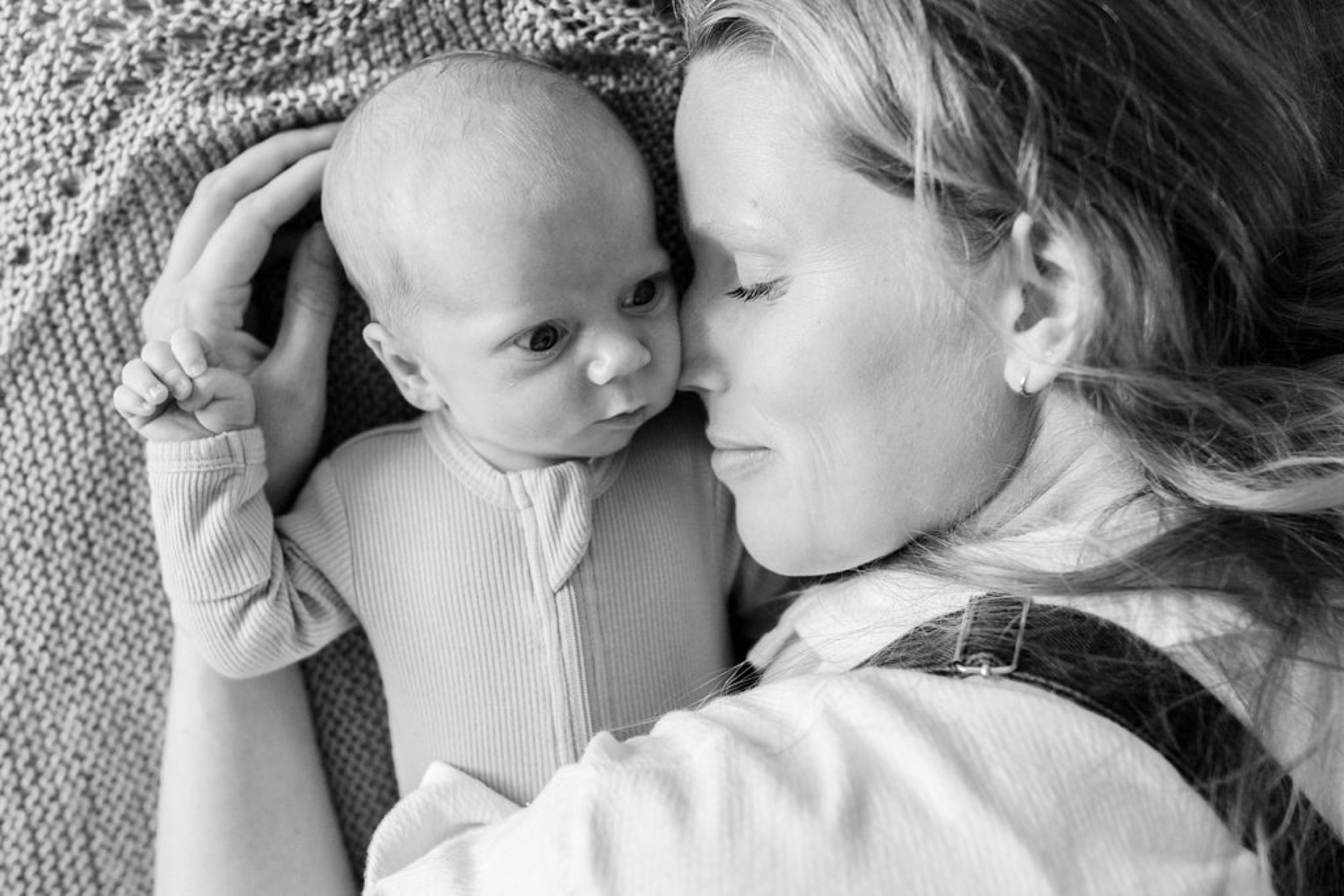 Mother gazes down at her newborn as the infant reaches up to touch her face, their connection intimate and tender on soft textured fabric