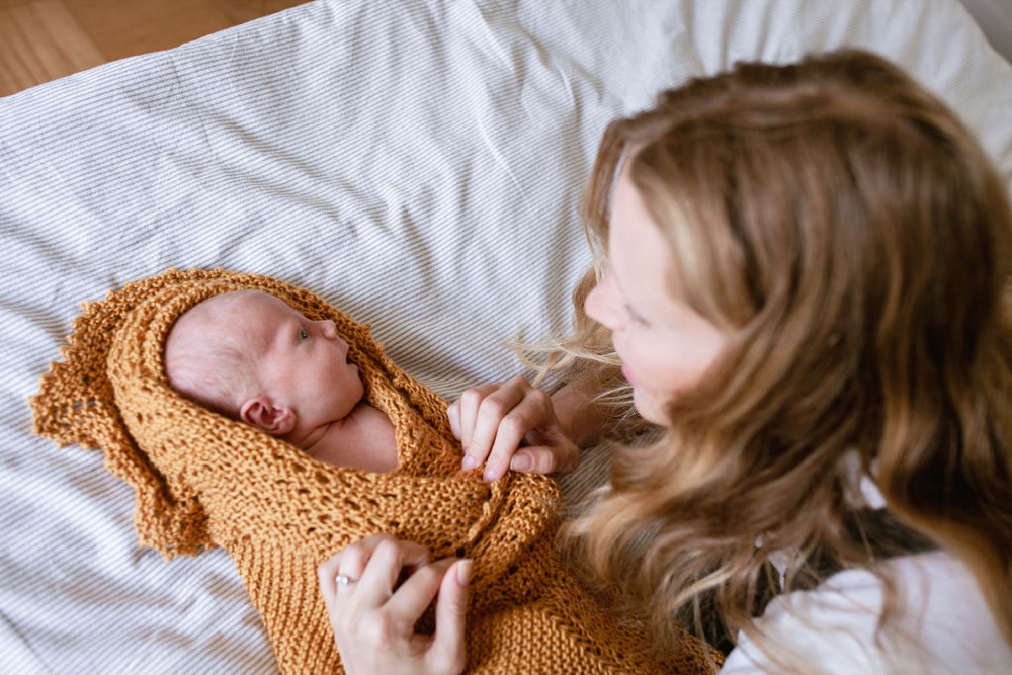 A toddler in a pink dress tenderly gazes at her newborn sibling swaddled in a golden knit wrap on white bedding