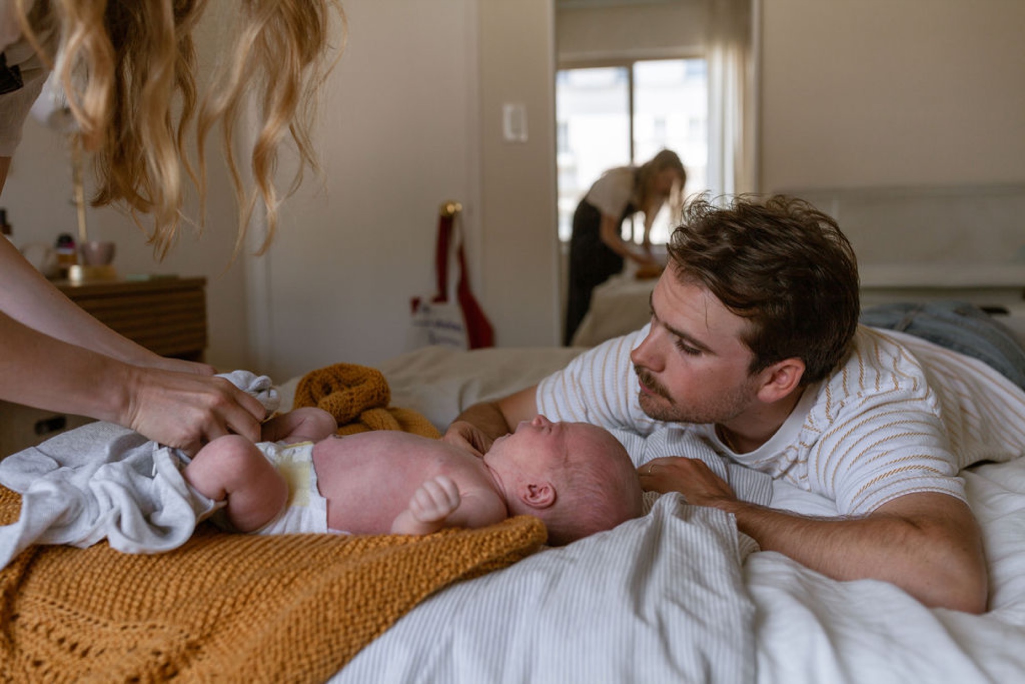 Father gazes tenderly at his newborn lying on a mustard blanket while the mother gently offers a stuffed bear during an intimate bedroom moment