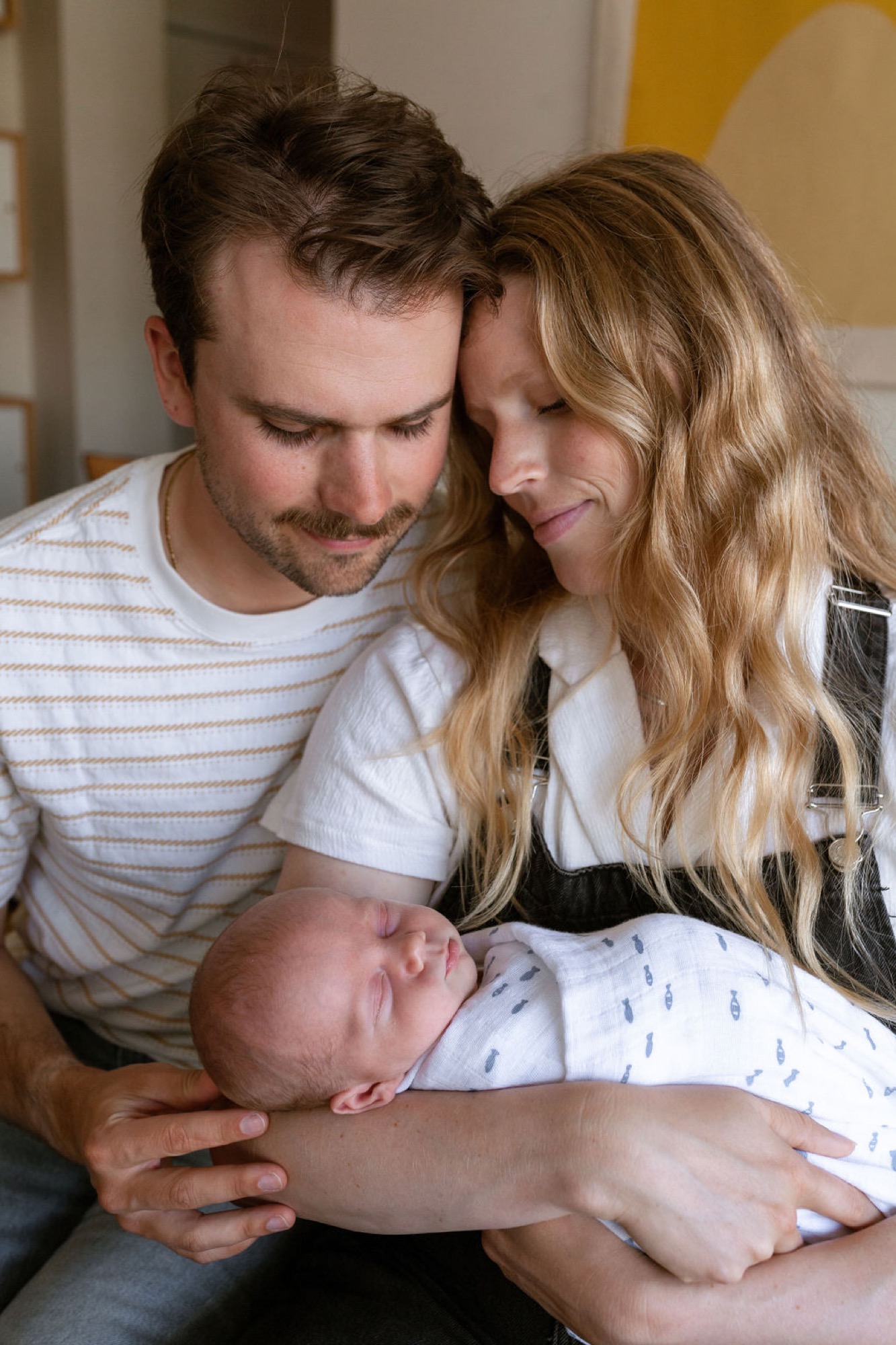 New parents gaze tenderly at their sleeping newborn, heads close together in warm indoor light