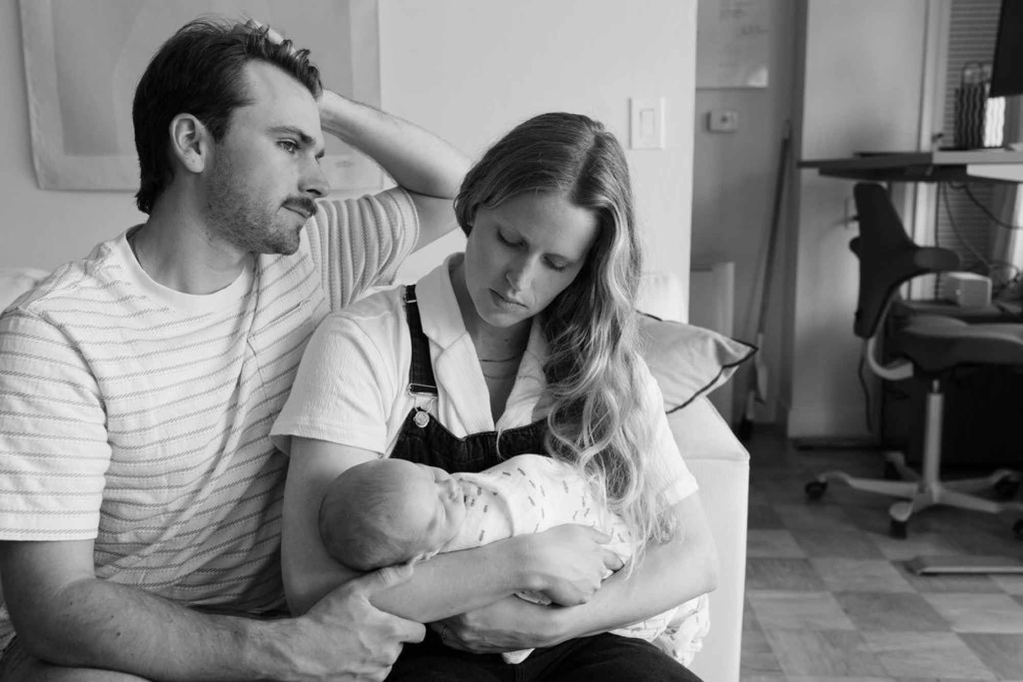 New parents gaze tenderly at their swaddled newborn in a bright home office, father's protective hand on mother's shoulder