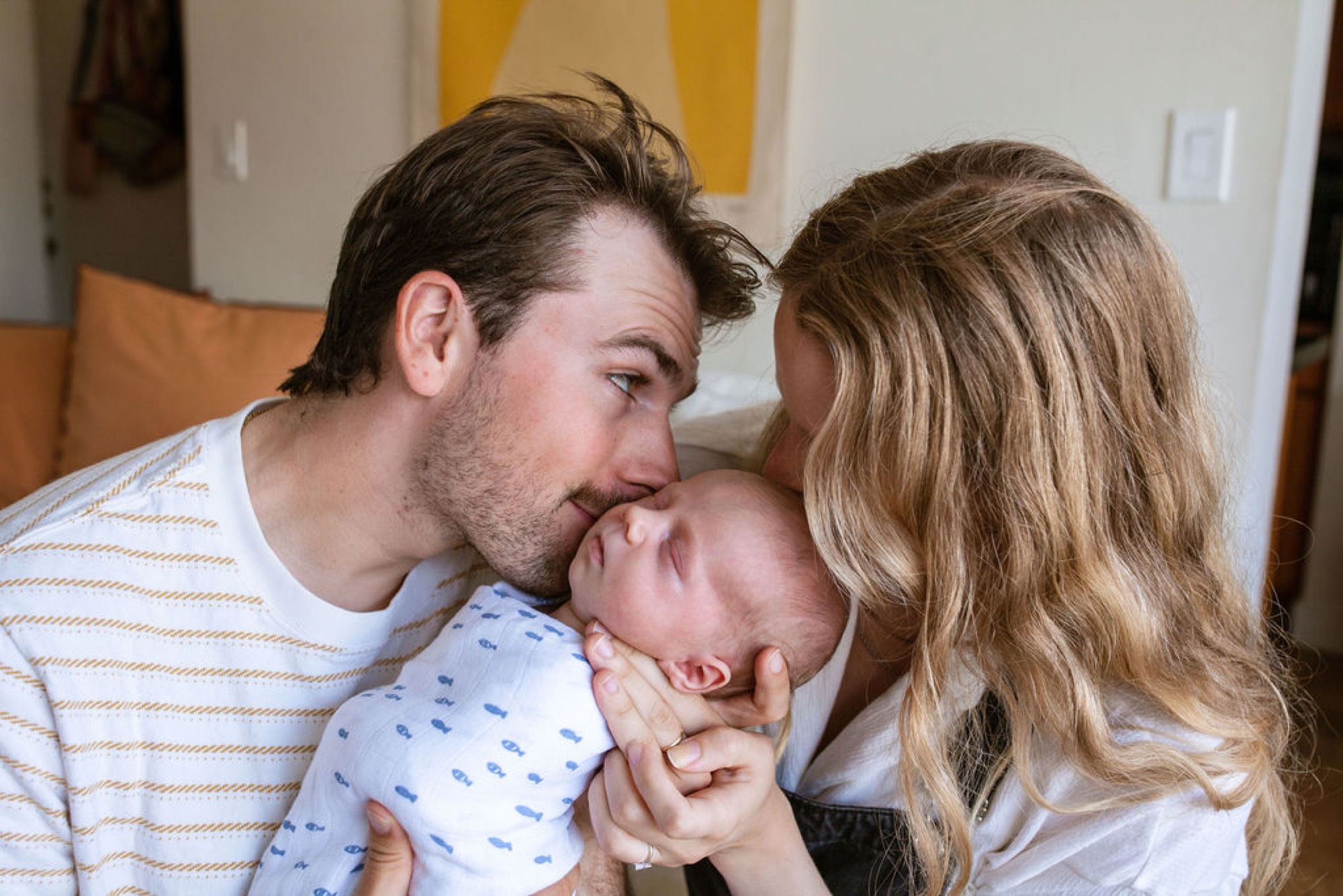 Father kisses newborn's forehead while mother gazes tenderly at their infant in warm natural light