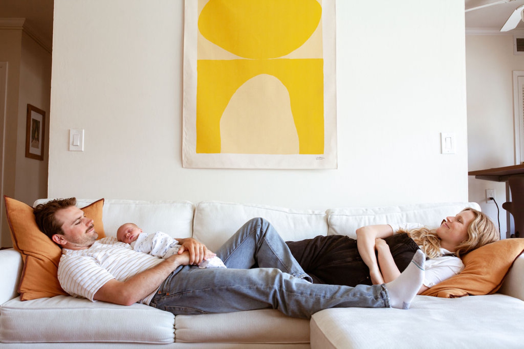 Father cradles newborn while mother relaxes beside them on a cream sofa beneath bright yellow abstract art