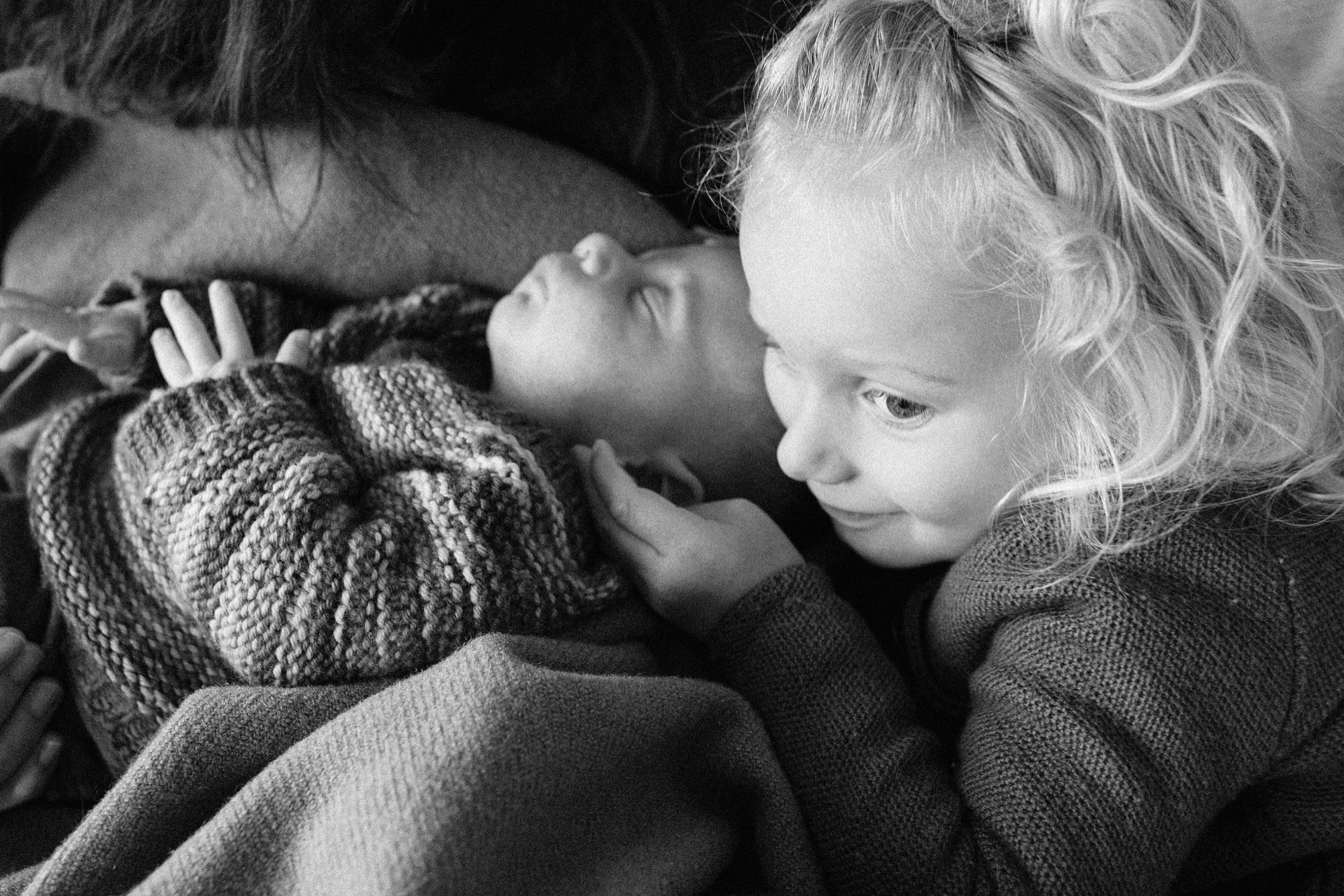 Two young siblings in soft knit sweaters gaze downward with tender curiosity, their profiles illuminated by gentle natural light in an intimate black and white portrait