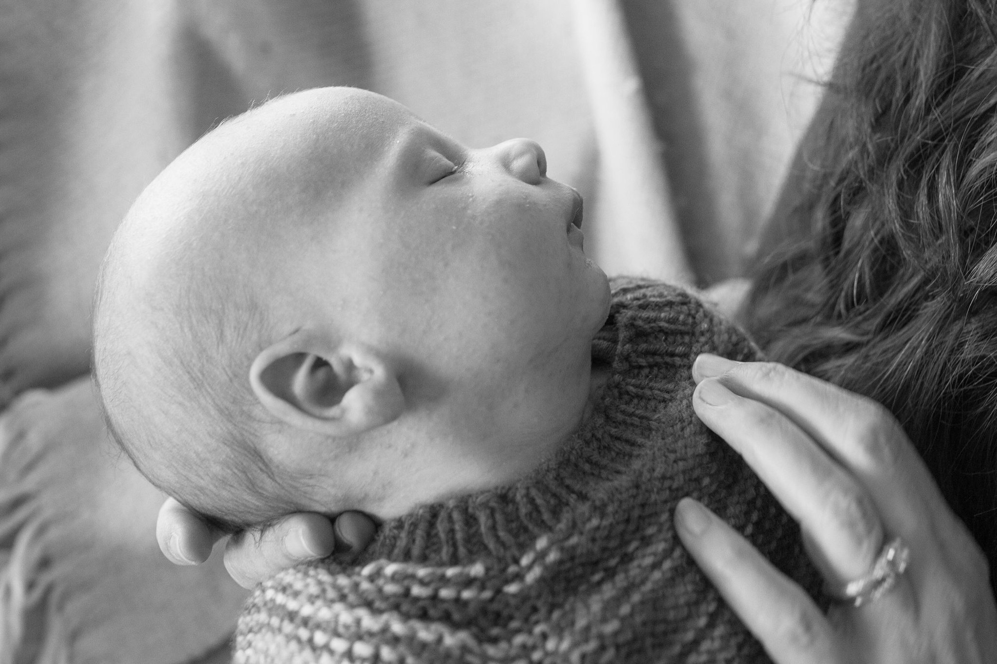 A newborn tilts their head back in peaceful contentment while cradled against a parent's shoulder in soft, diffused indoor light