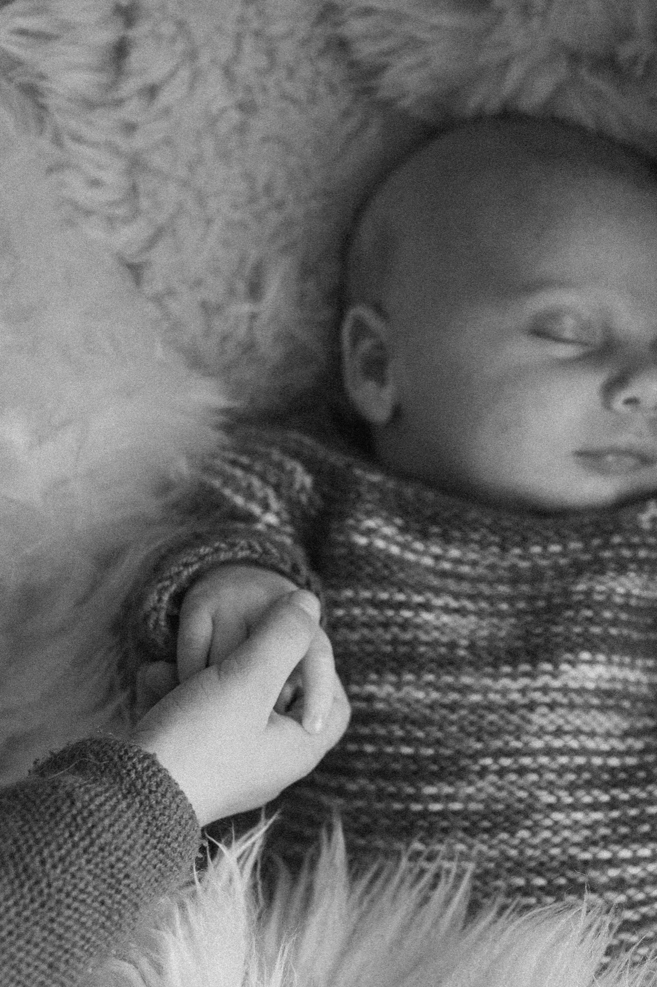 Newborn's delicate hand grasps parent's sweater as soft, diffused light illuminates their profile in intimate repose