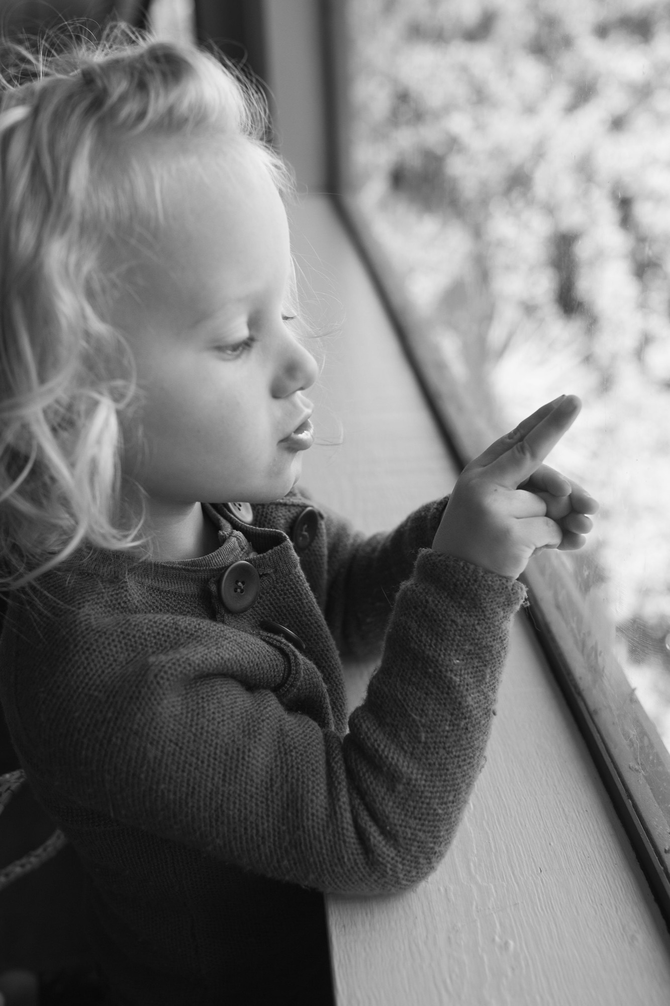 Toddler in knit cardigan gazes thoughtfully at snowy window, small hand pressed against cold glass