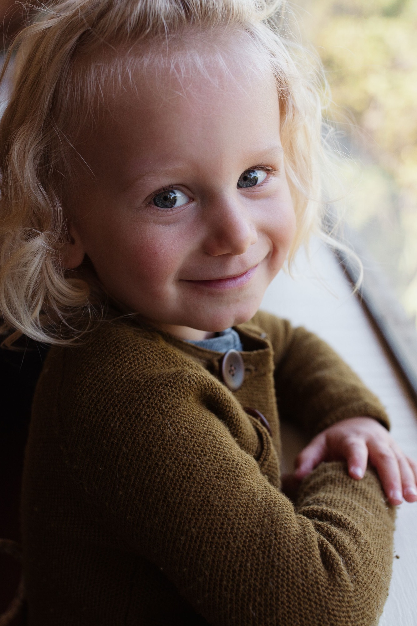 Toddler in mustard cardigan gazes over shoulder with genuine smile, soft natural light catching blonde curls and bright eyes