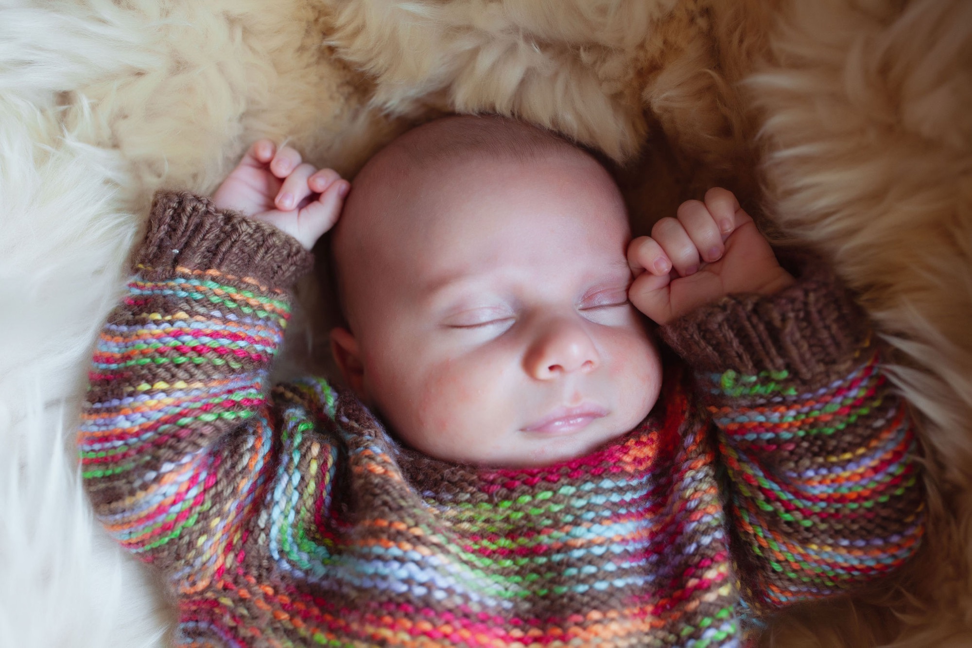 Peacefully sleeping newborn swaddled in a vibrant multicolored knit blanket against soft cream fur