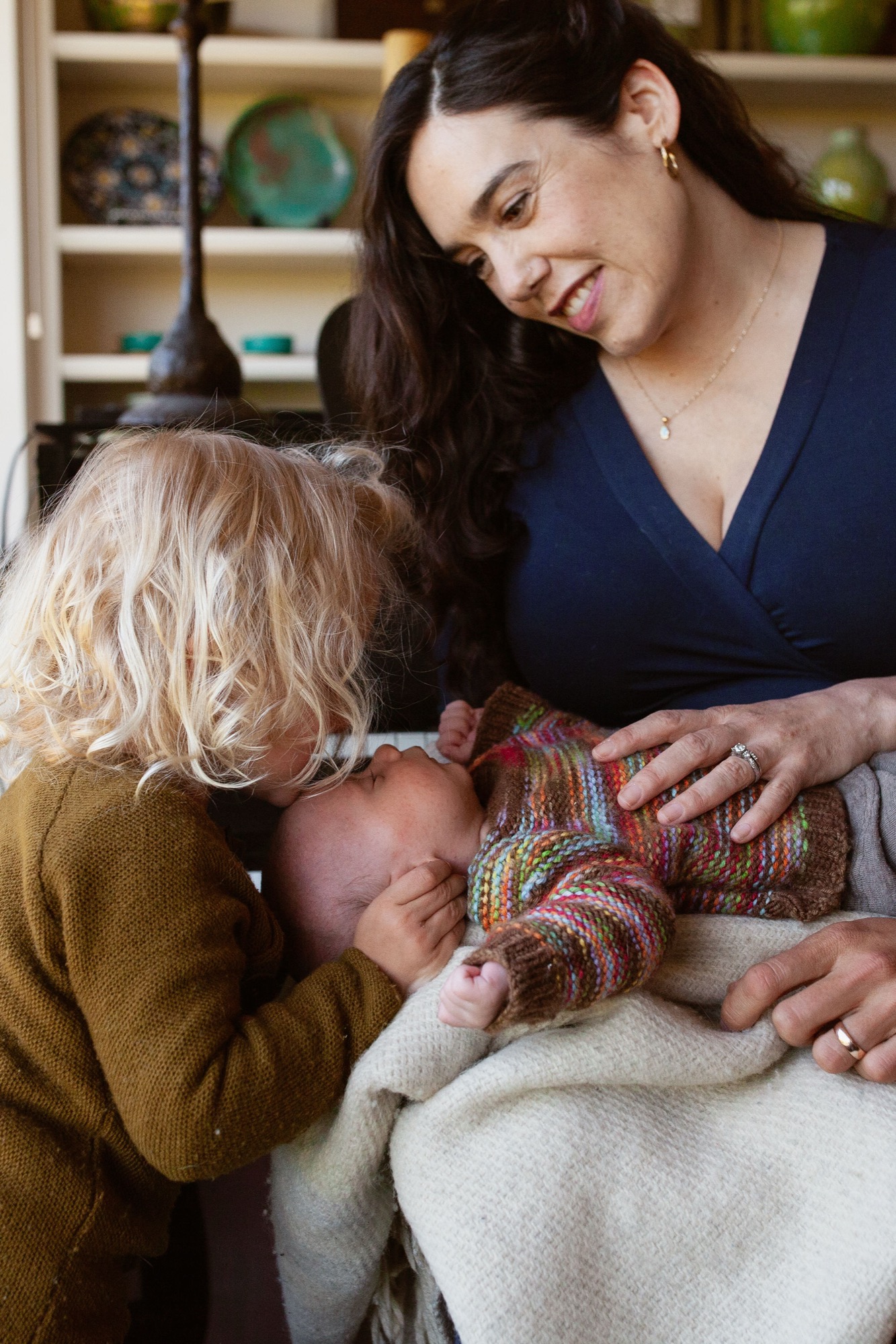 A toddler in mustard yellow leans close to examine a newborn's face while the mother smiles down at both children in warm indoor light