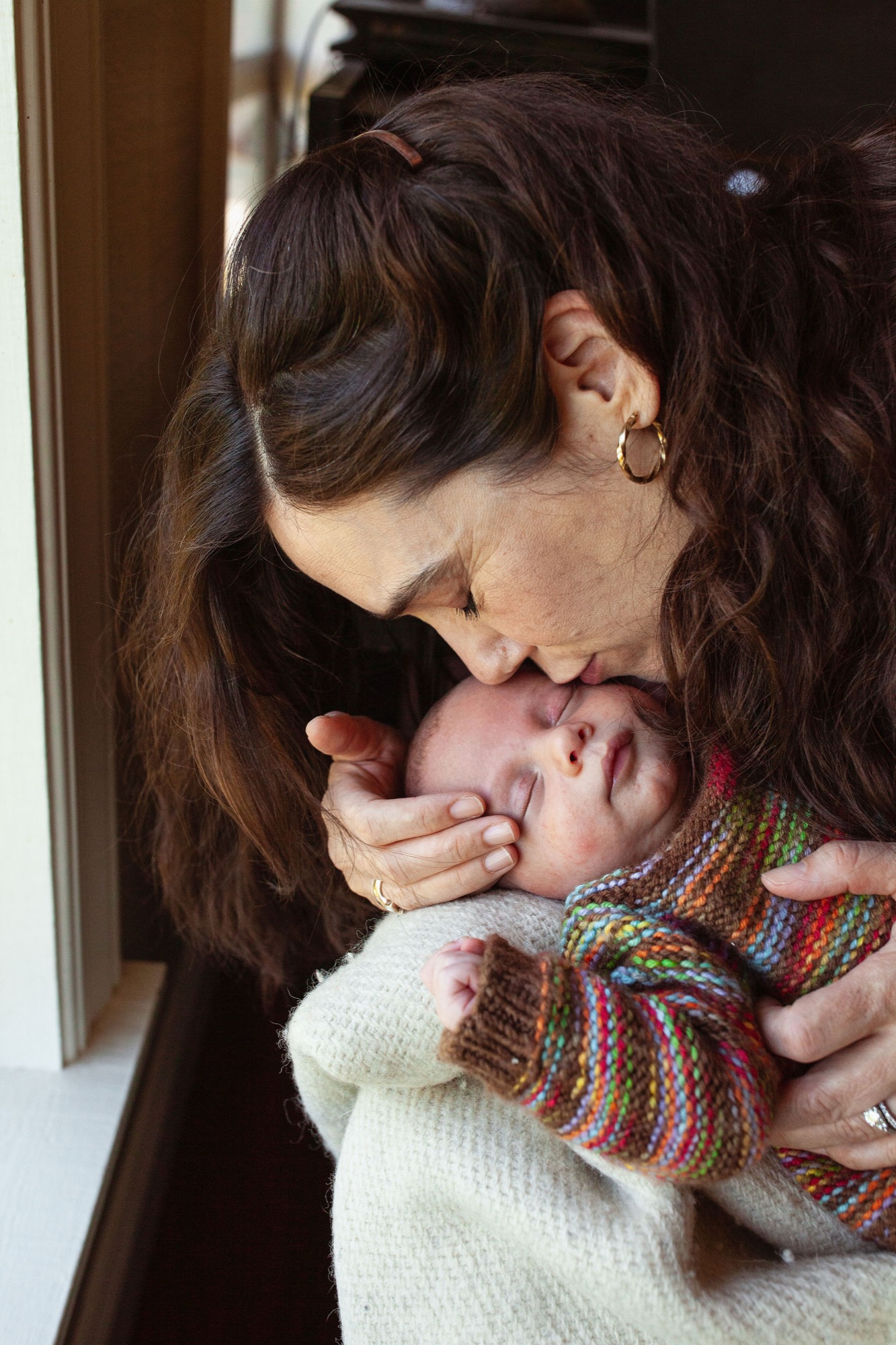 Mother gazes tenderly at newborn wrapped in colorful striped sweater, bathed in soft window light