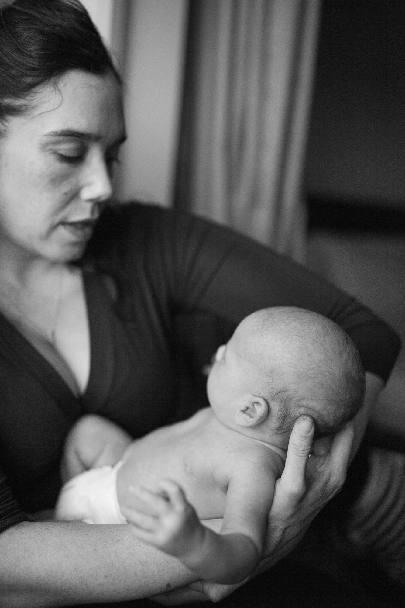 Mother gazes tenderly at newborn cradled in her hands, soft window light illuminating their intimate profile against dark fabric