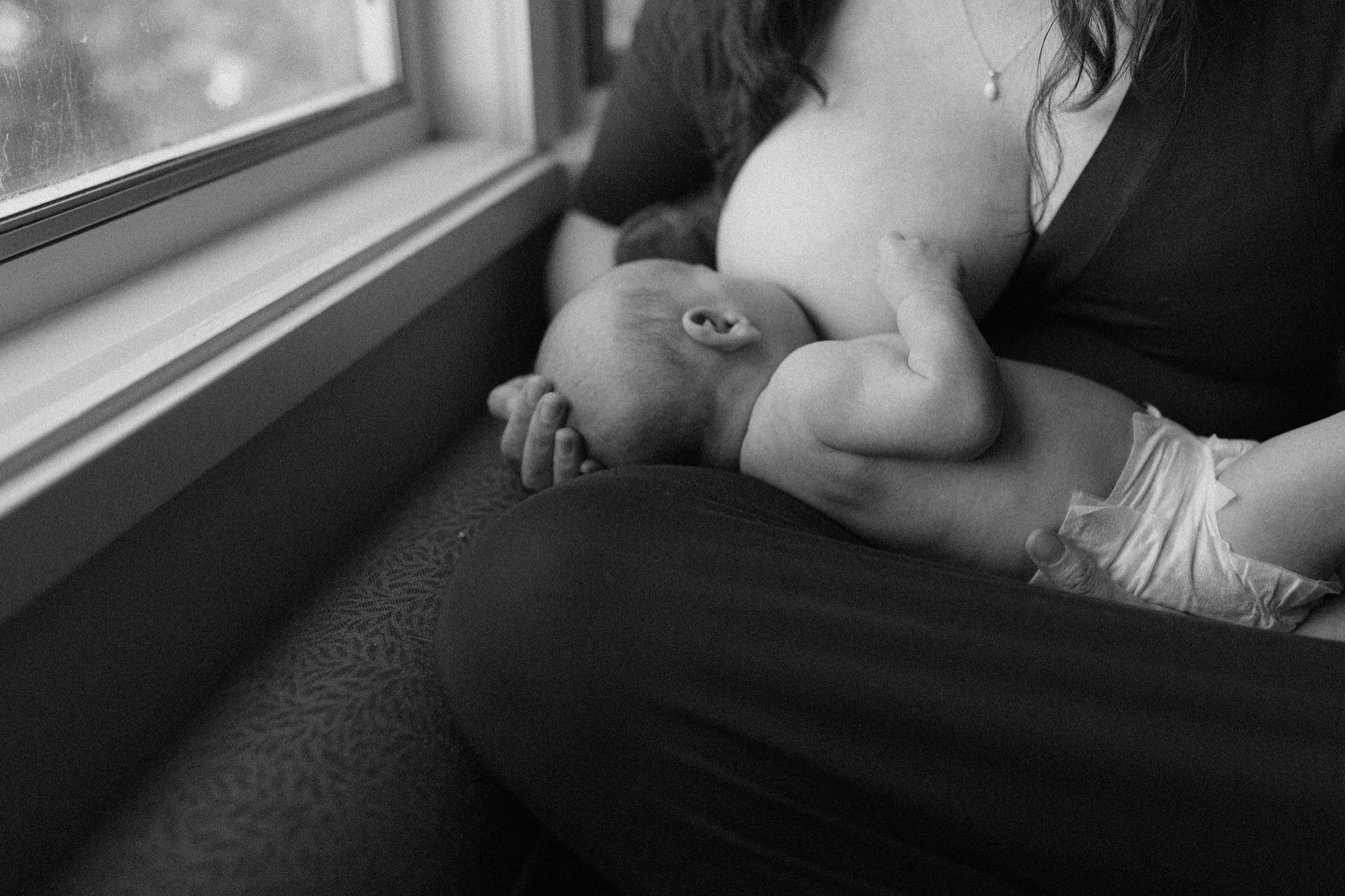 A newborn sleeps peacefully in mother's arms by a sunlit window, her gentle gaze downward capturing profound tenderness