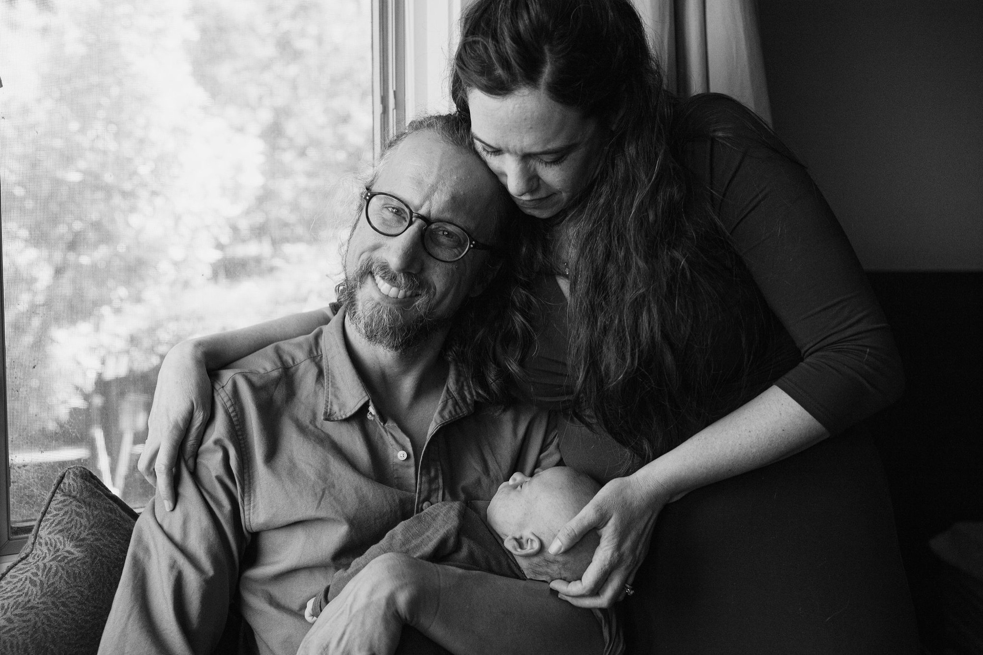 A bearded father in glasses gazes at his newborn while the mother leans in close by the bright window, their tender expressions radiating quiet joy
