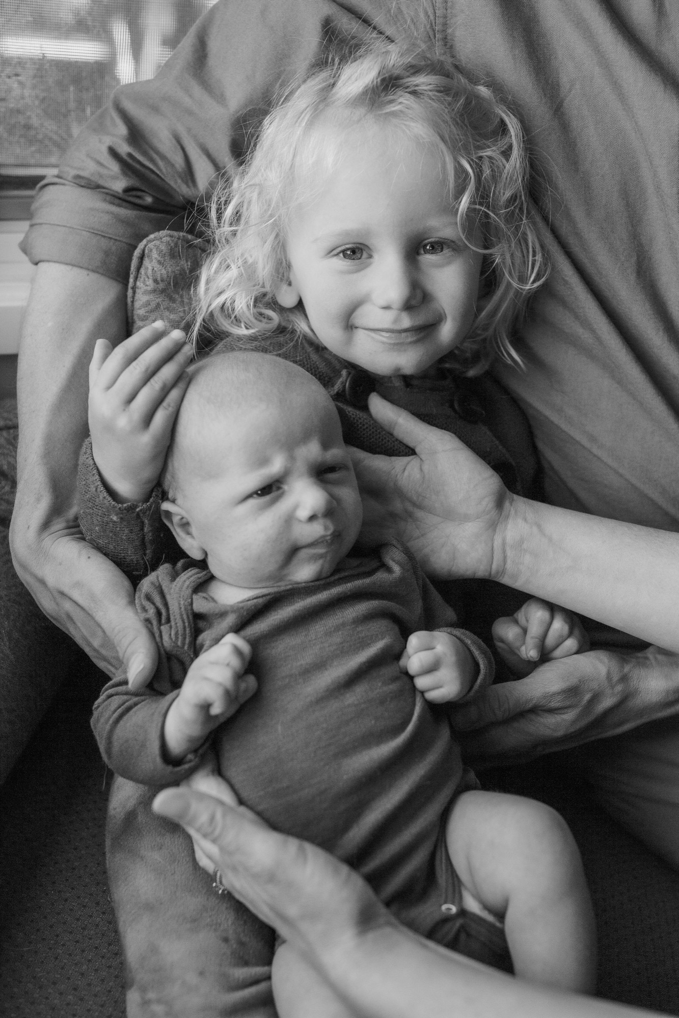 Older sister beams at camera while cradling newborn brother, their hands intertwined in gentle embrace