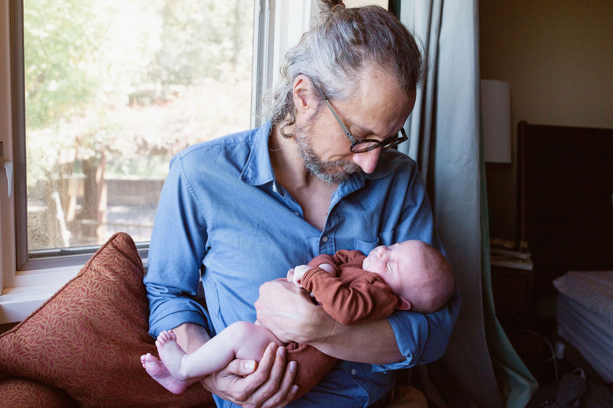 A bearded man in glasses holds his newborn by the window, gazing down with tender affection as natural light floods the room