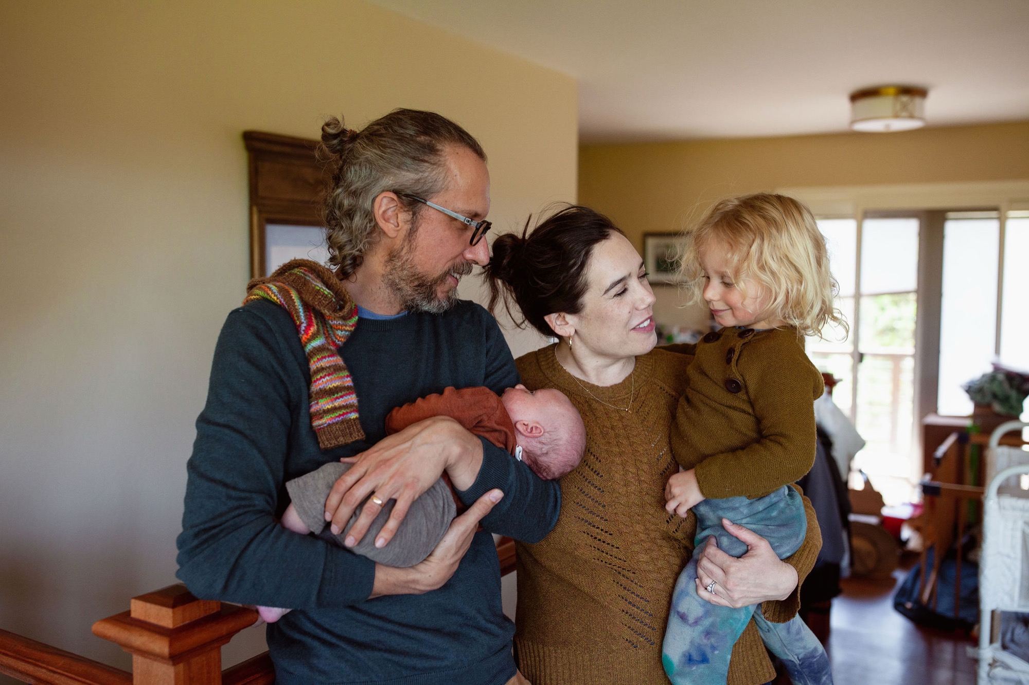 A father holds his newborn while the mother cradles their toddler, all four smiling warmly in golden afternoon light streaming through home windows