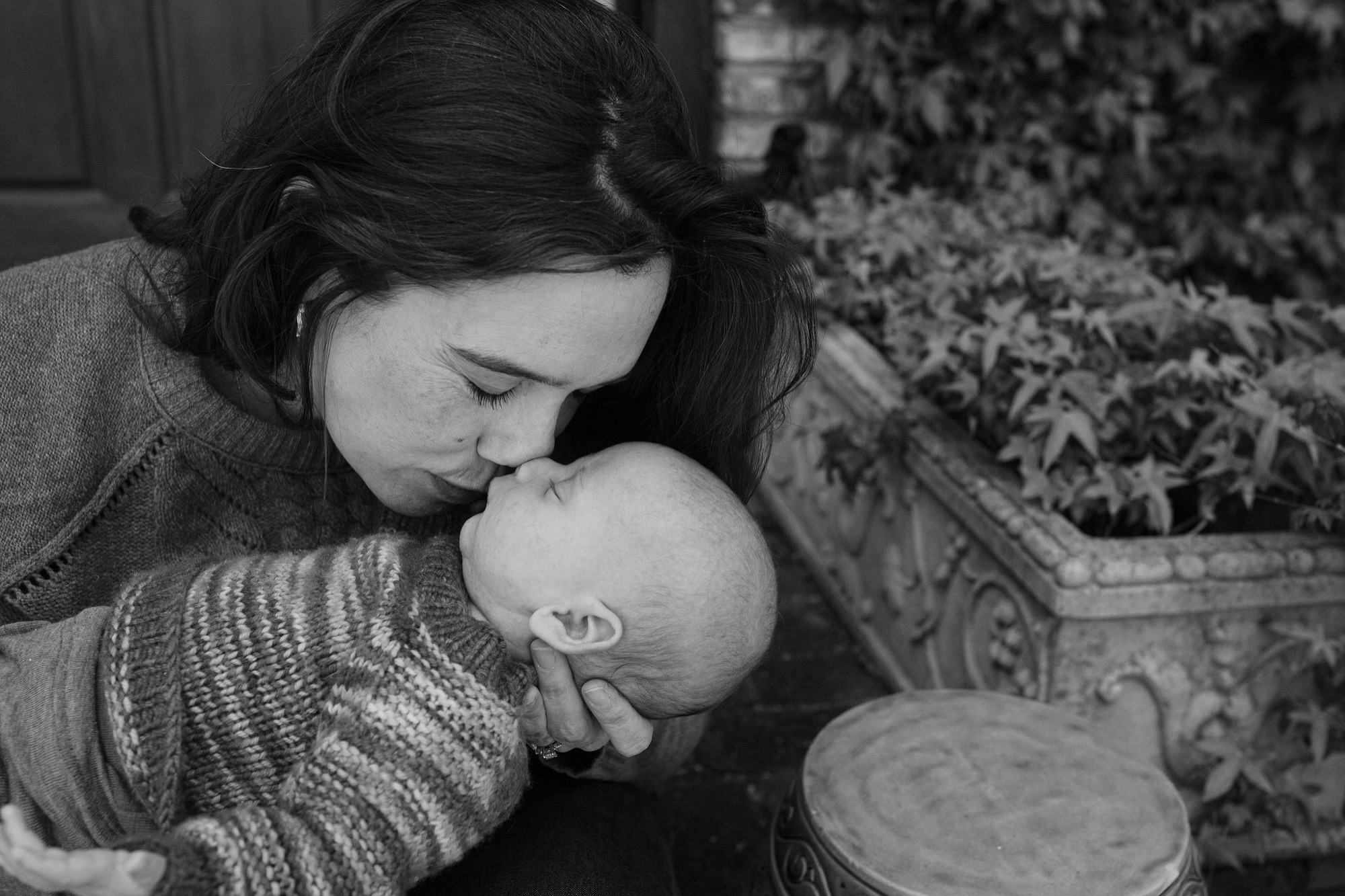 A mother tenderly kisses her newborn's head in soft natural light, her dark hair framing their intimate moment indoors