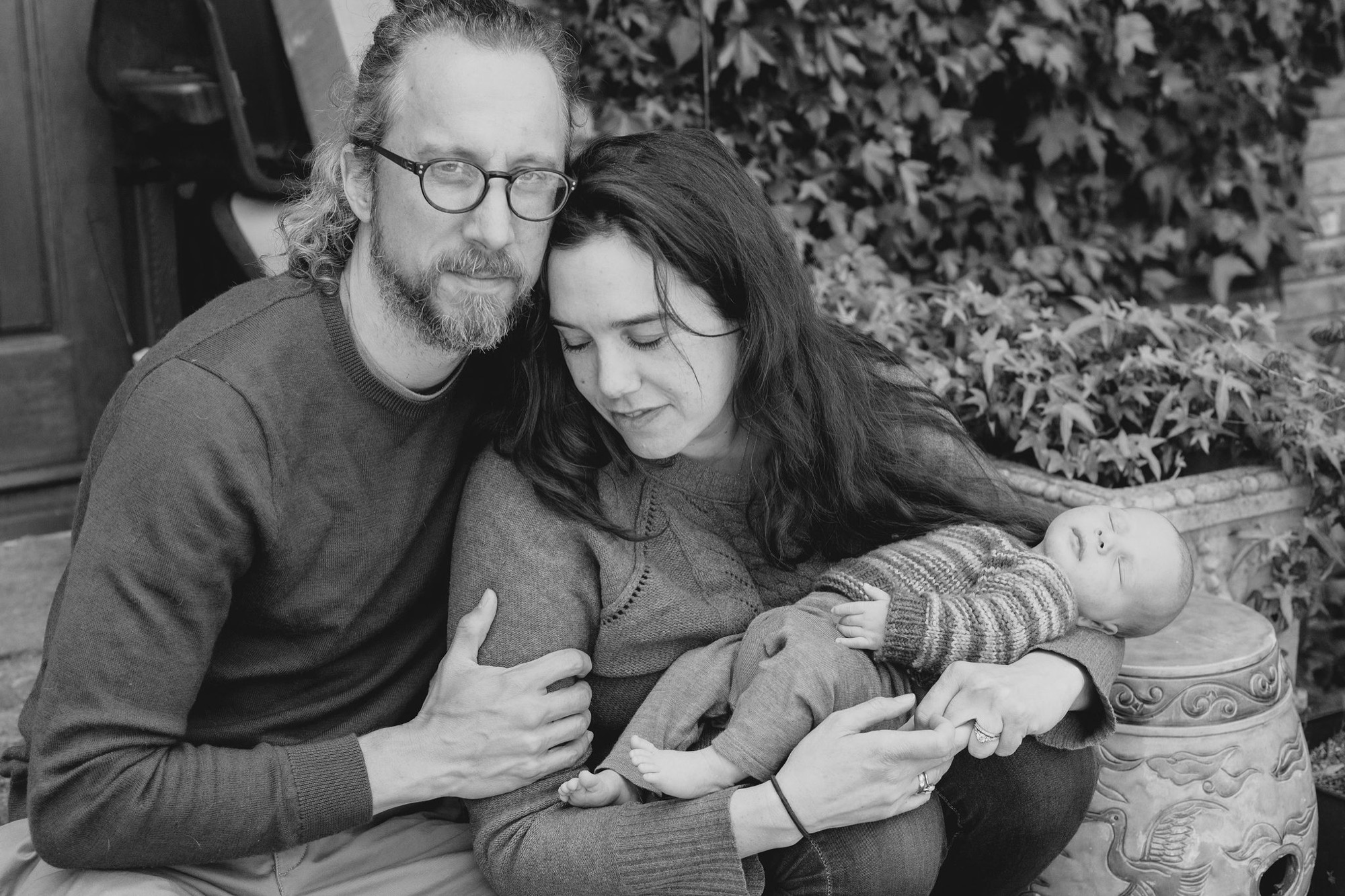 Parents gaze tenderly at their newborn cradled between them beside ivy-covered walls, soft natural light illuminating their gentle expressions