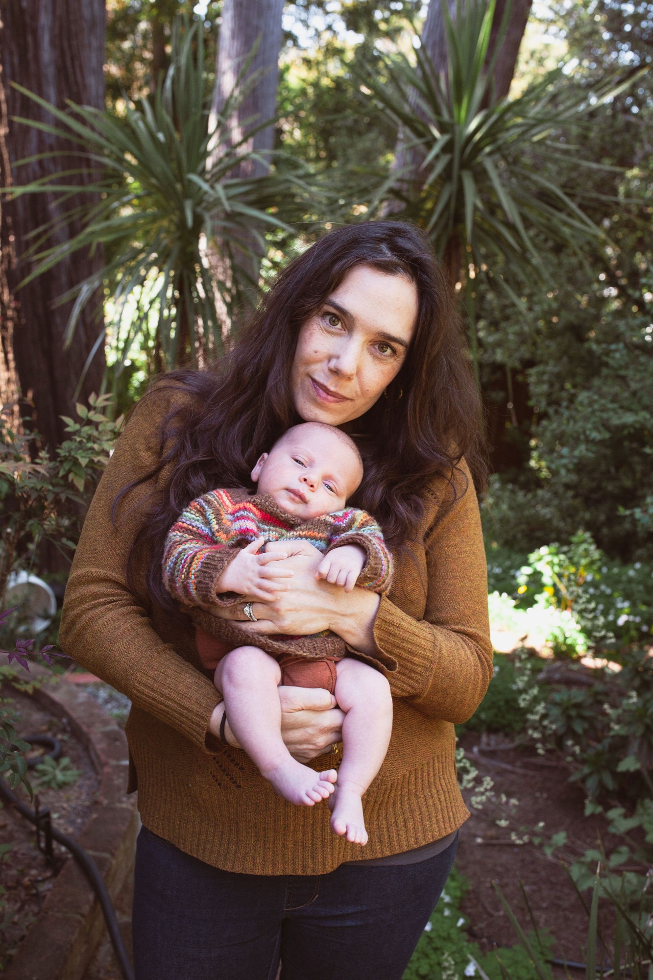 Mother gazes warmly at camera while cradling newborn in colorful sweater among redwoods, dappled sunlight filtering through canopy