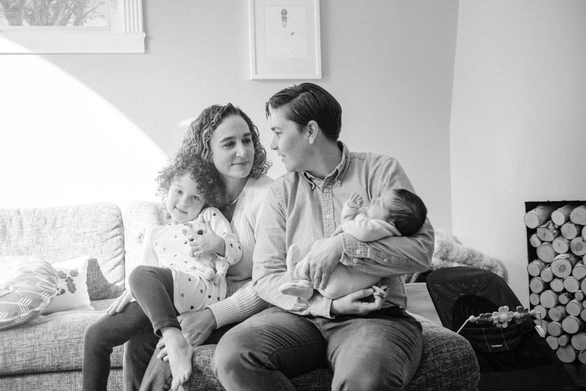 Two moms sit together on a cream linen sofa, one gazing at the other while she smiles gently, their two young children nestled between them in warm natural light