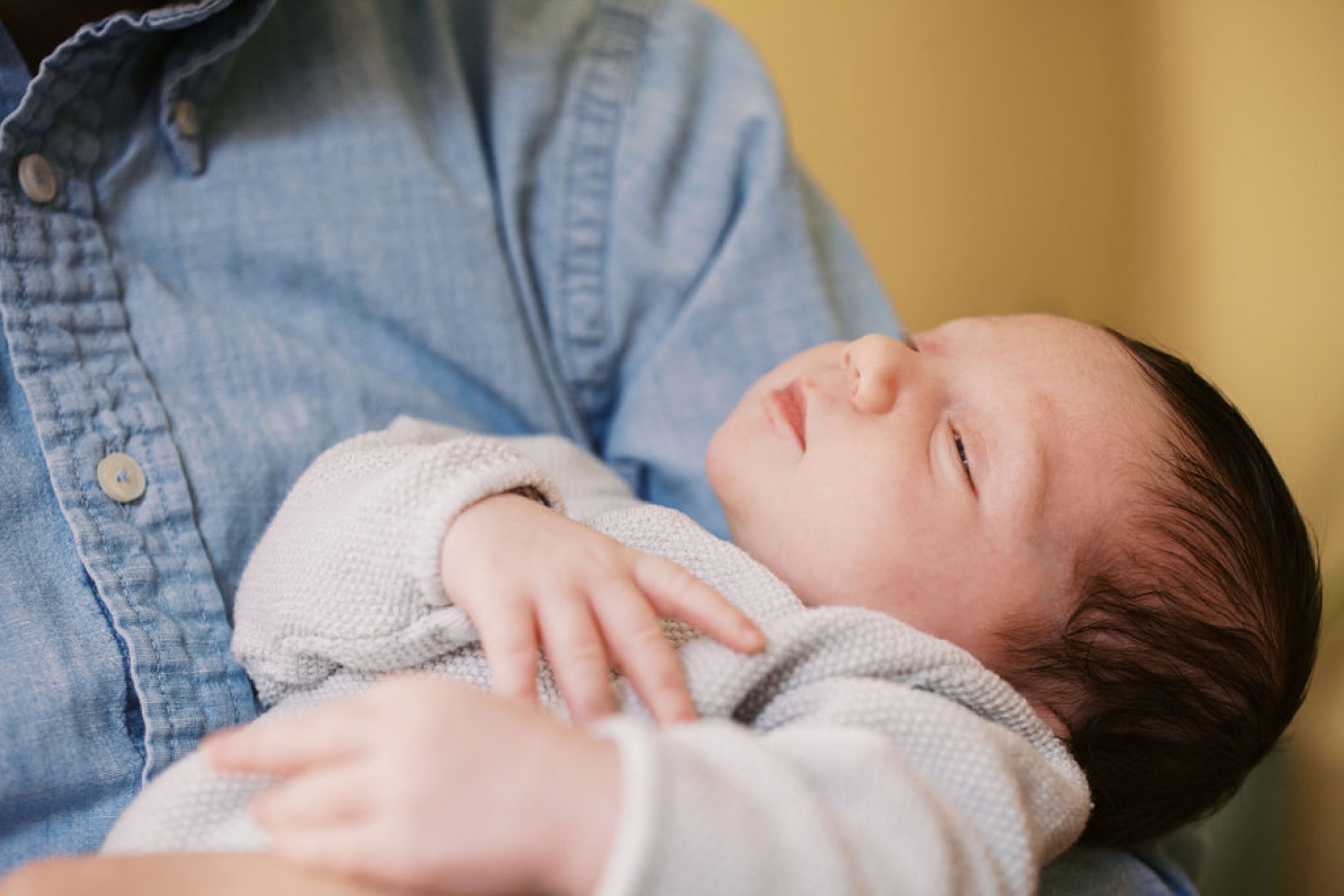 Peacefully swaddled newborn sleeps in parent's arms wearing a cream knit blanket against a soft yellow wall