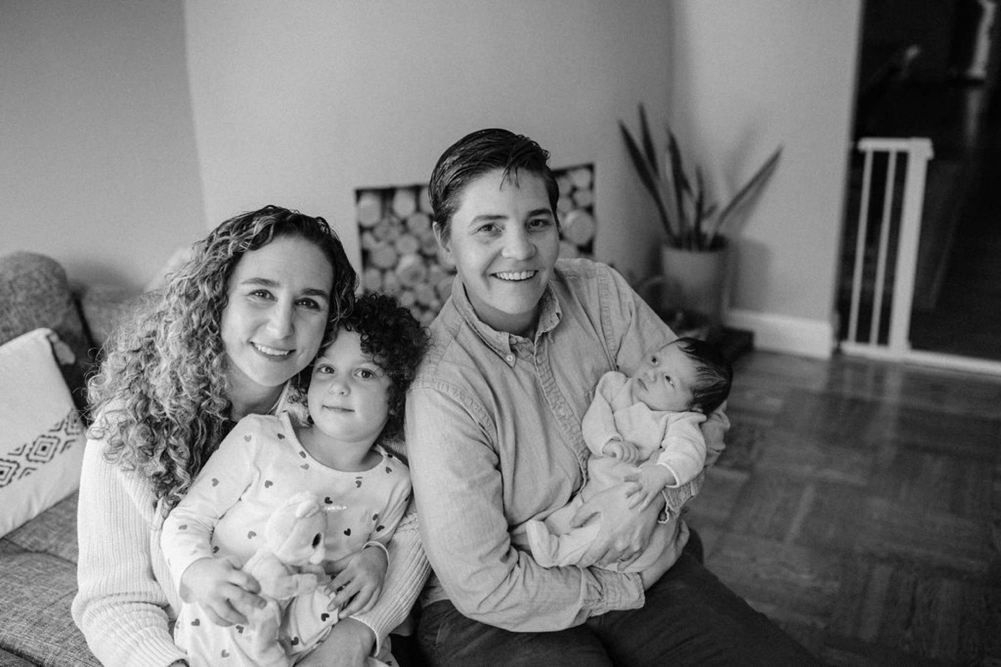 A joyful family of four sits together on a living room floor, the parents beaming while holding their newborn and toddler in natural window light