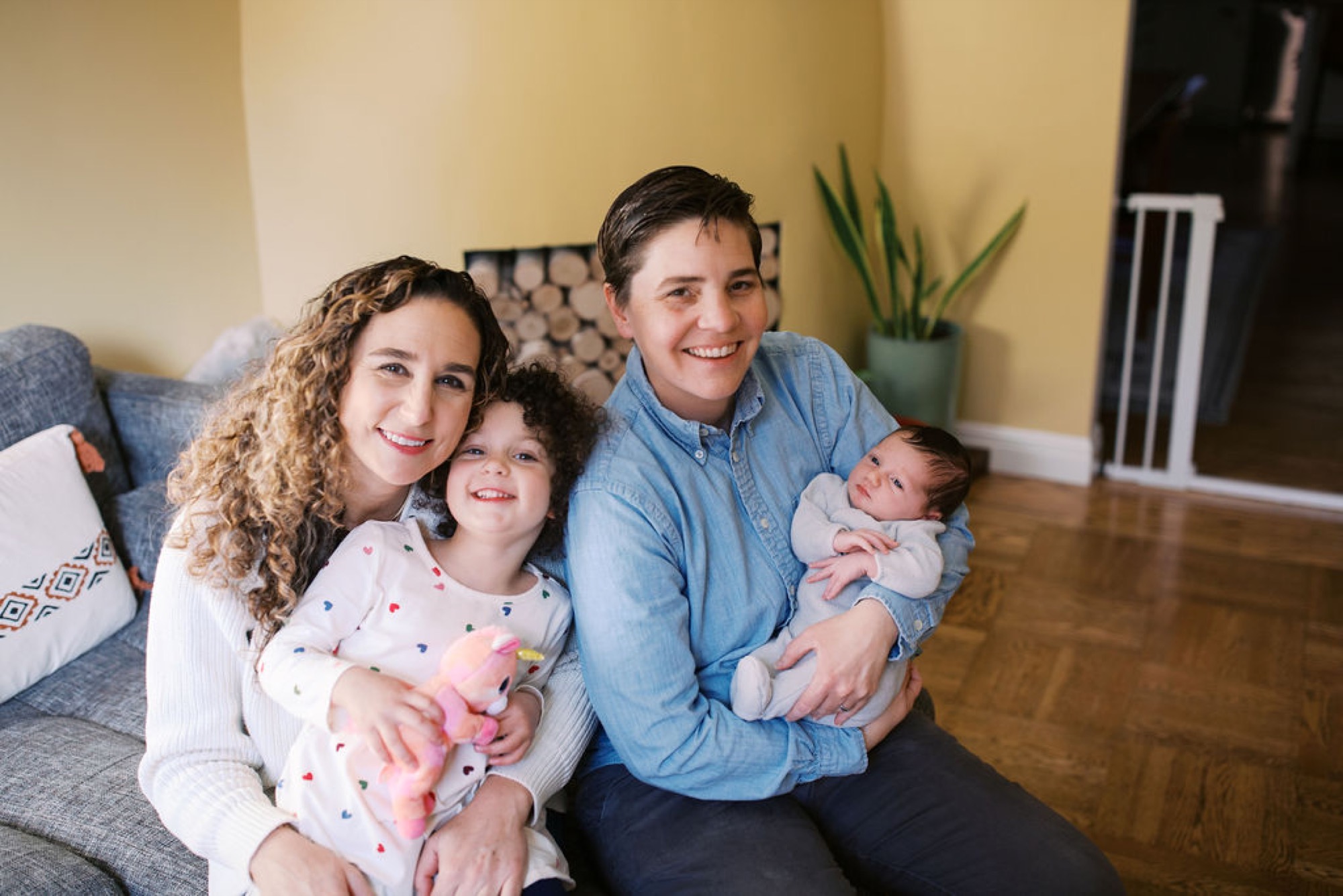 A beaming family of four sits close together on a gray couch, with warm yellow walls and natural light illuminating their genuine smiles and relaxed togetherness