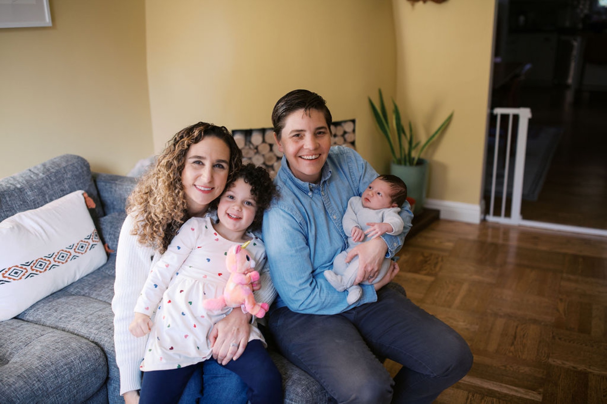 Young family of four beams at camera while seated on gray couch, warm yellow walls and natural light creating intimate living room portrait