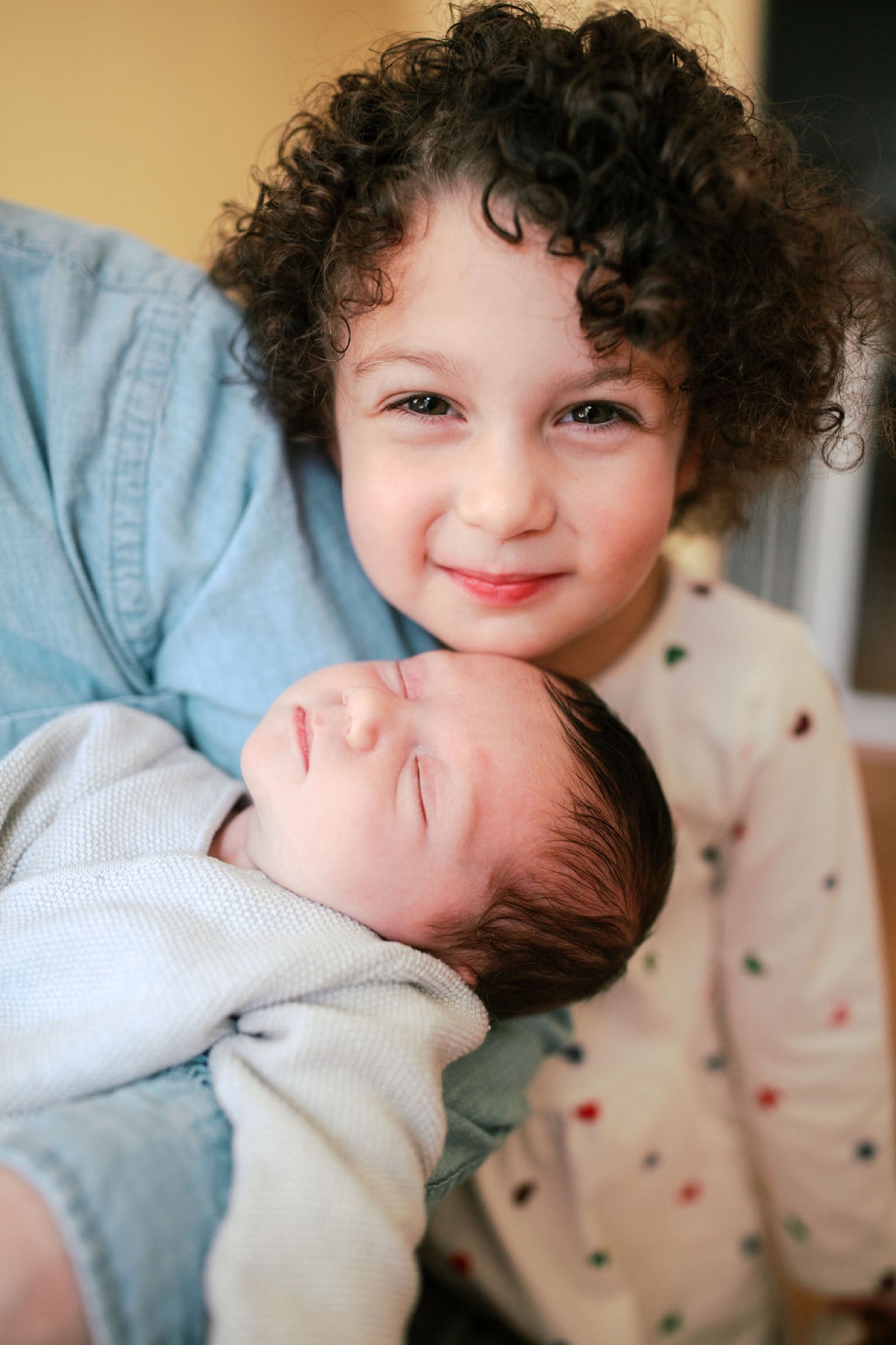 Toddler with dark curly hair gazes warmly at camera while cradling newborn sibling in blue denim shirt indoors