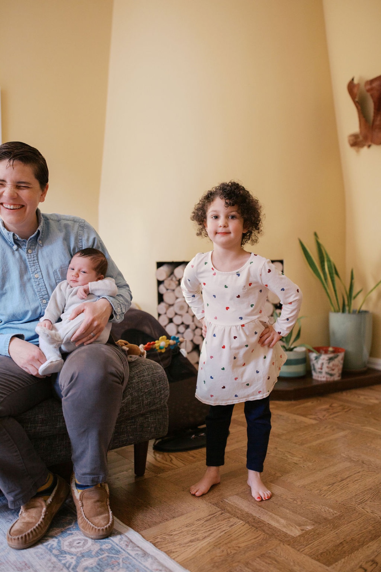 A smiling parent holds their infant while their older daughter with curly hair stands barefoot beside them in a warm, sunlit living room