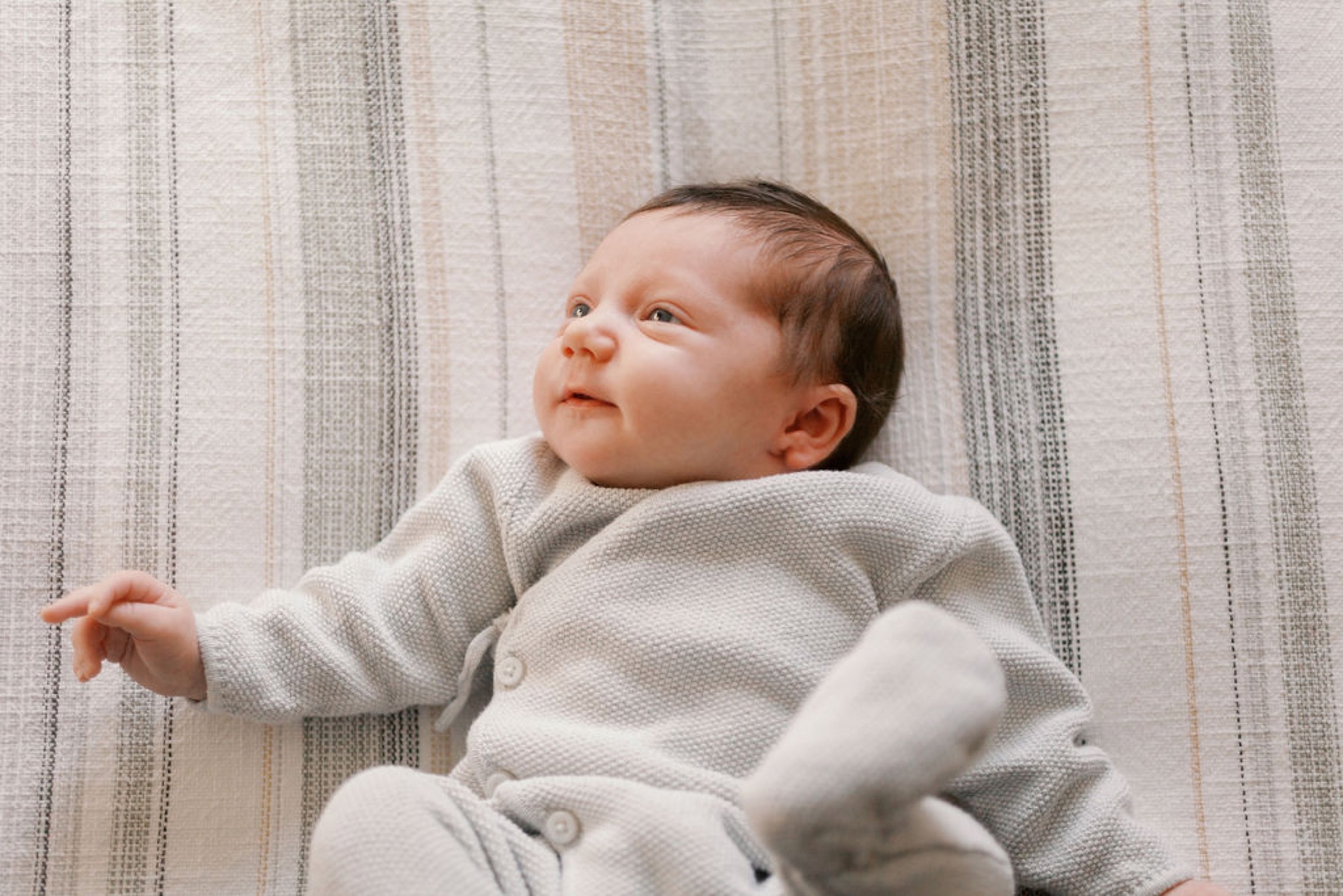 A newborn in cream waffle-knit clothing gazes thoughtfully upward, one arm extended, against a soft striped linen backdrop