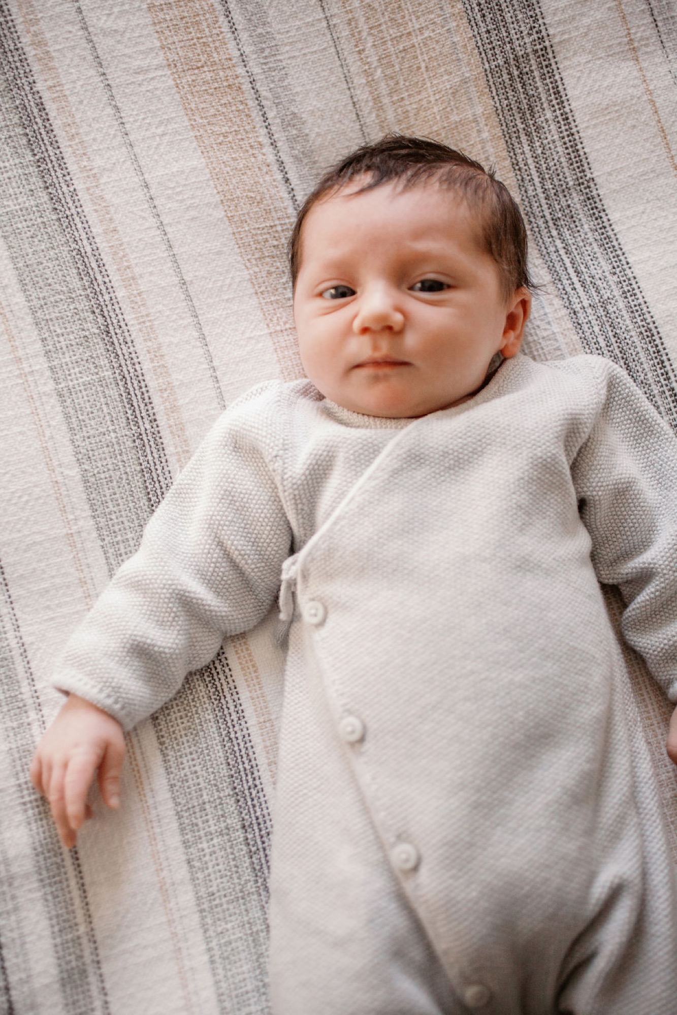 Newborn swaddled in cream knit wrap lies on striped neutral bedding, gazing directly at camera with alert, serious expression