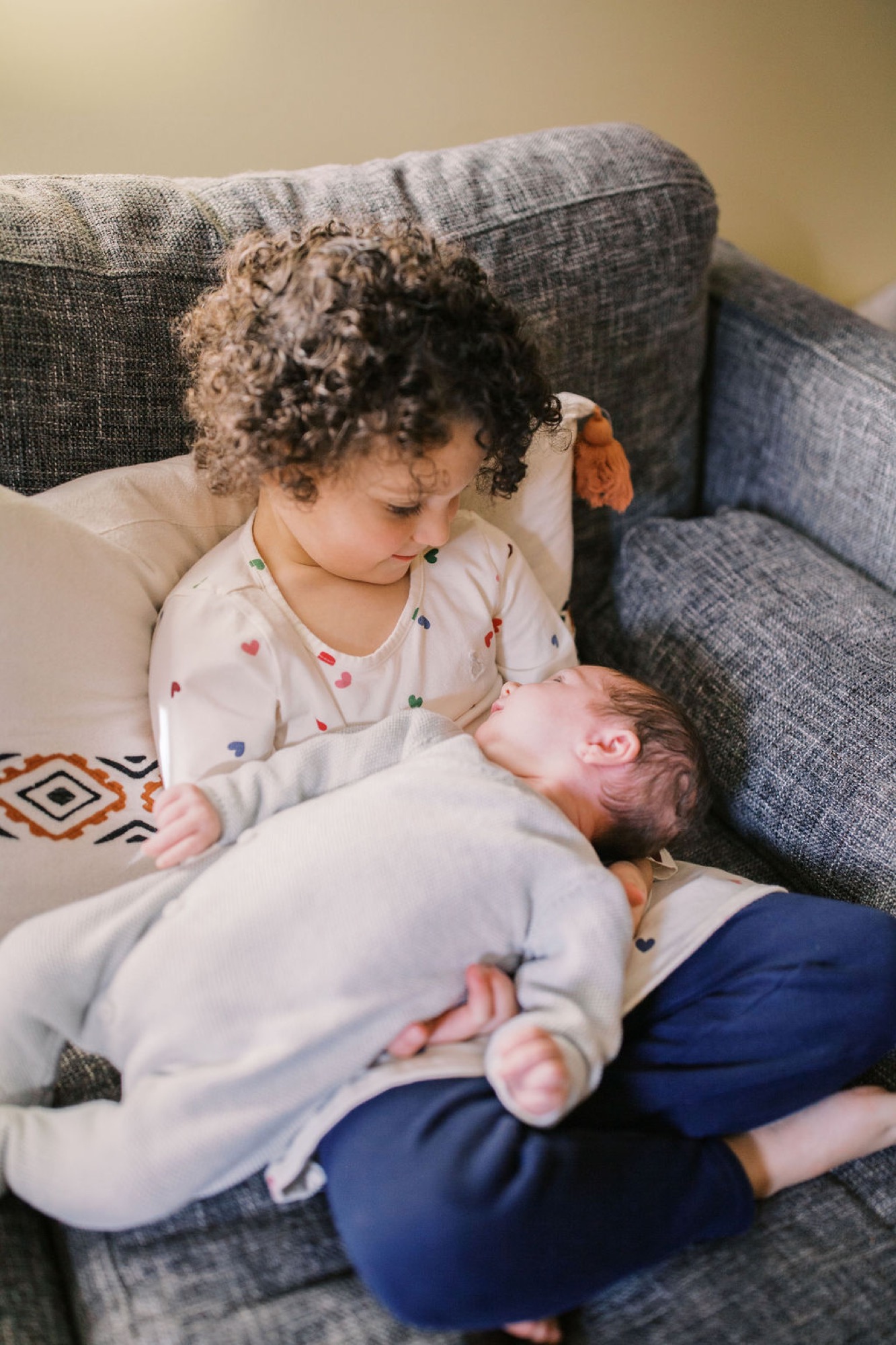 Toddler in heart-patterned shirt gazes tenderly at newborn sibling on gray couch, soft natural light illuminating their gentle connection